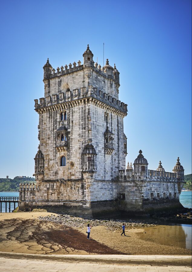Belém Tower in Lisbon stands tall against a clear blue sky, with two people walking on the sandy shore below.