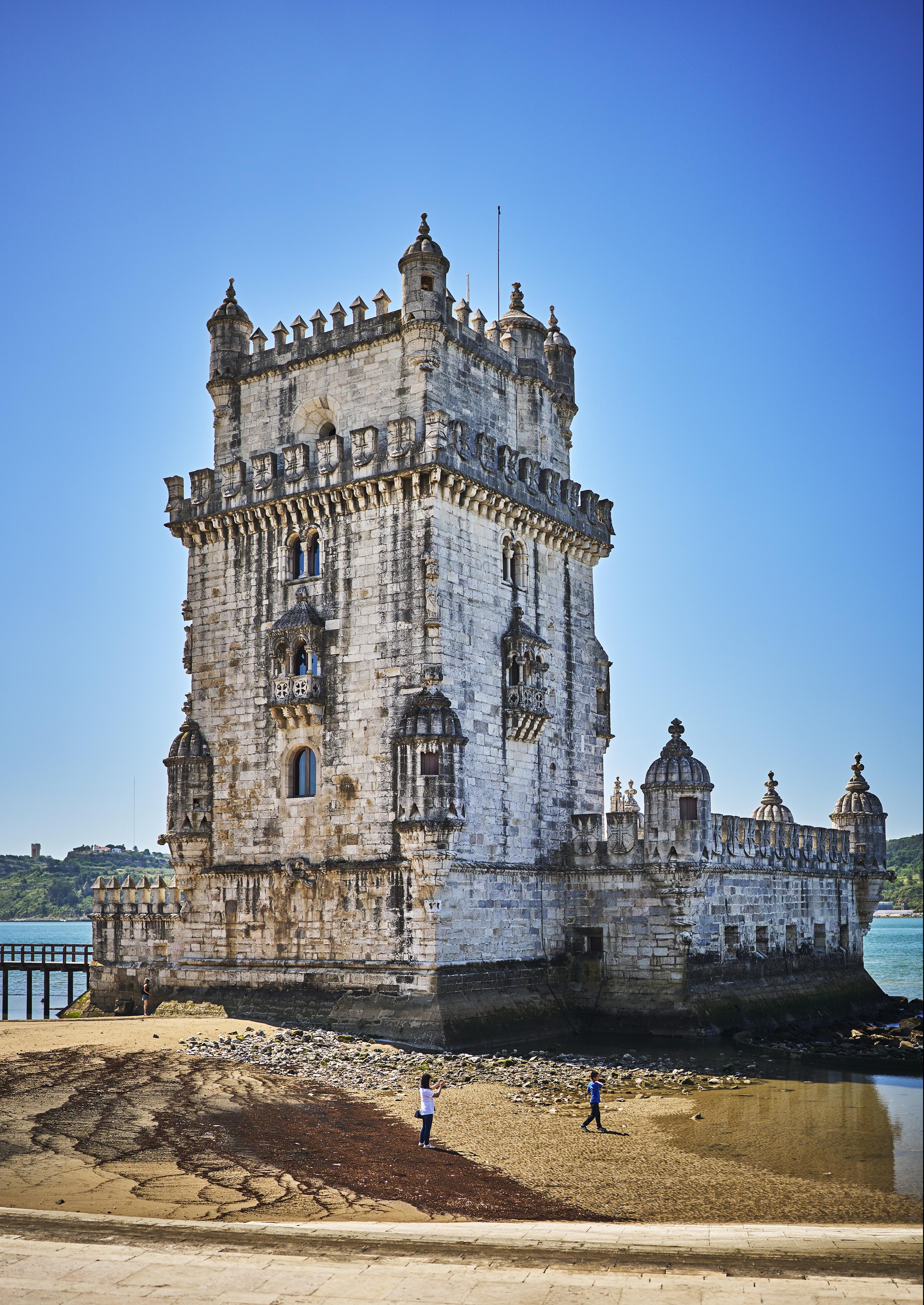 Belém Tower in Lisbon stands tall against a clear blue sky, with two people walking on the sandy shore below.