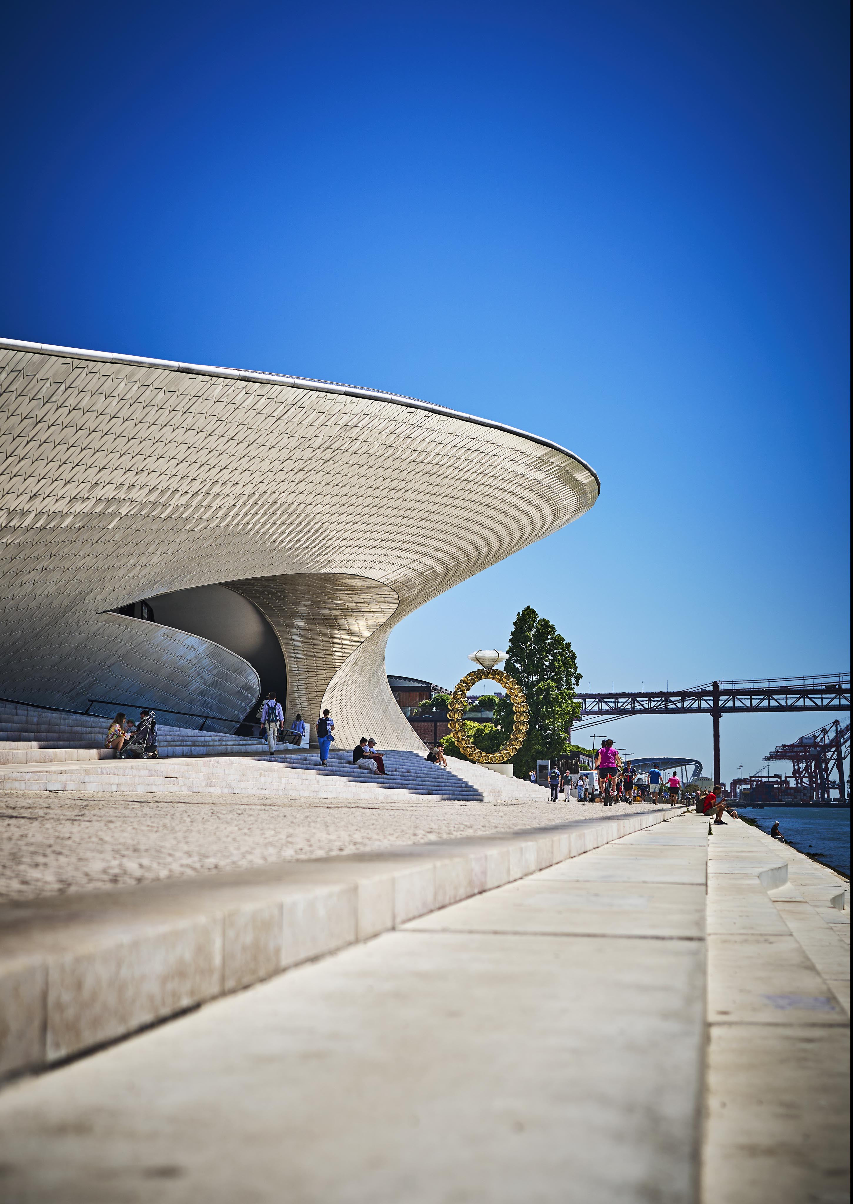 Modern architectural building with a curved design beside a waterfront, set against a clear blue sky with a bridge in the background and people walking nearby.