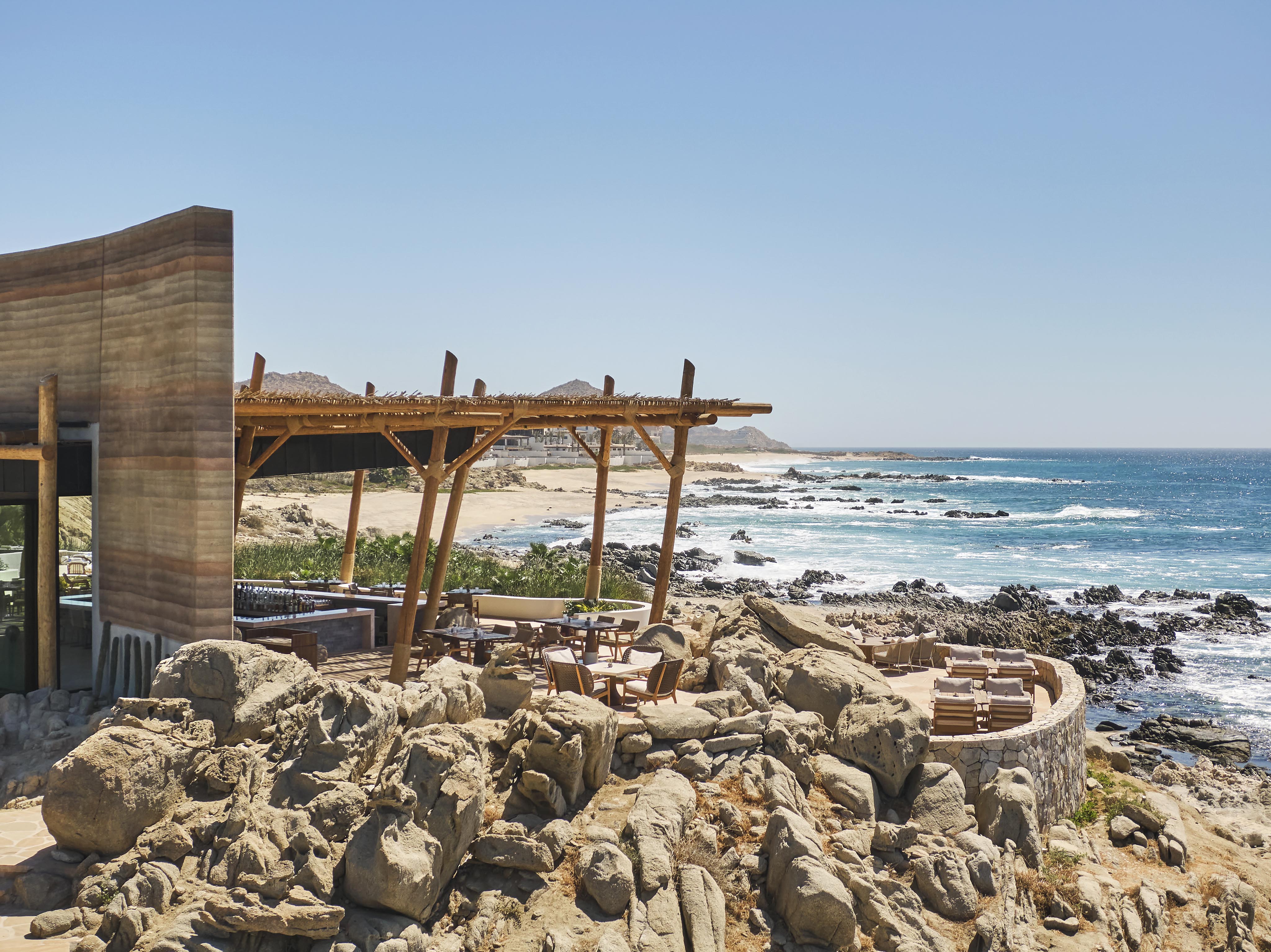 Seaside restaurant with Cove Club Cabo Del Sol contemporary modern structure overlooking a rocky coastline, blue sky, and waves in the background.