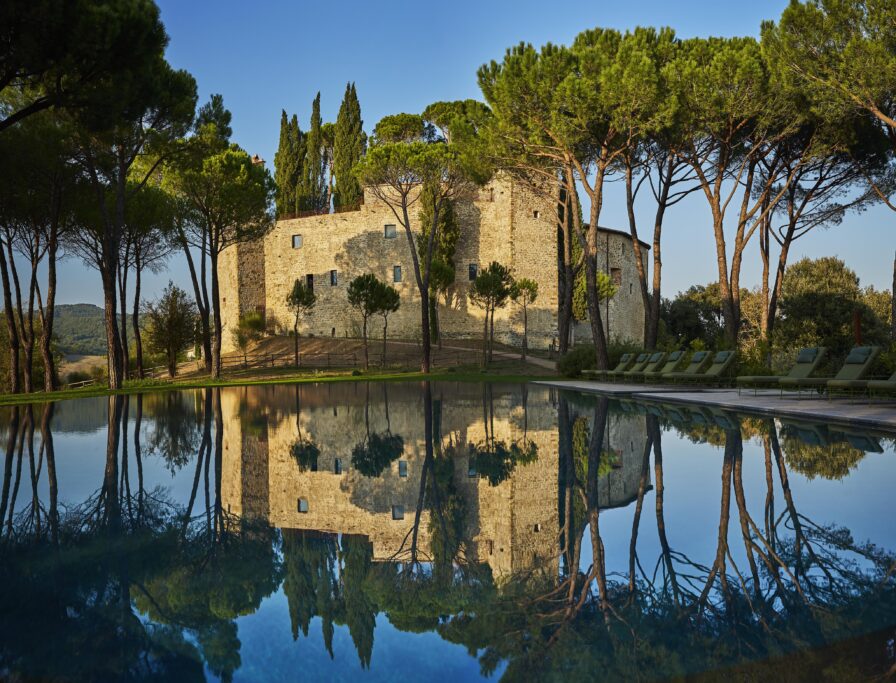 Historic stone building surrounded by trees, reflected in a large, tranquil pool under a clear blue sky.
