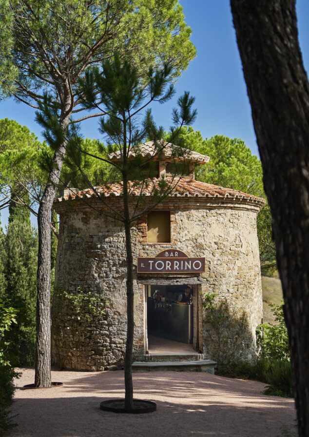 Stone building with a round structure, labeled "Bar Il Torrino," surrounded by tall trees under a clear blue sky.