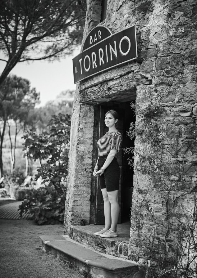 Woman standing in a doorway of a rustic stone building with a sign reading "Bar Il Torrino" above the entrance. Trees and plants in the background. Black and white photo.