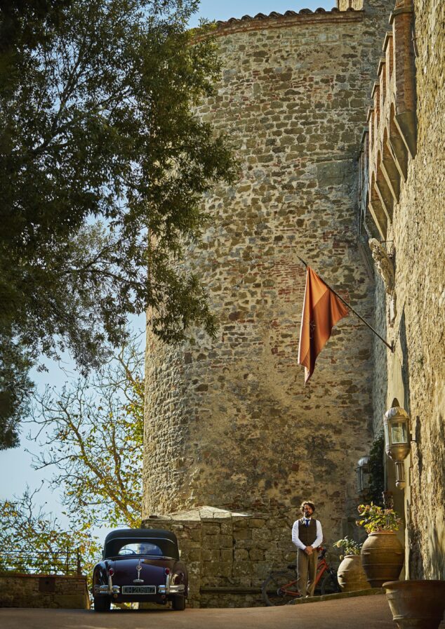 A man stands next to a vintage car and bicycle by a large stone building with a flag.