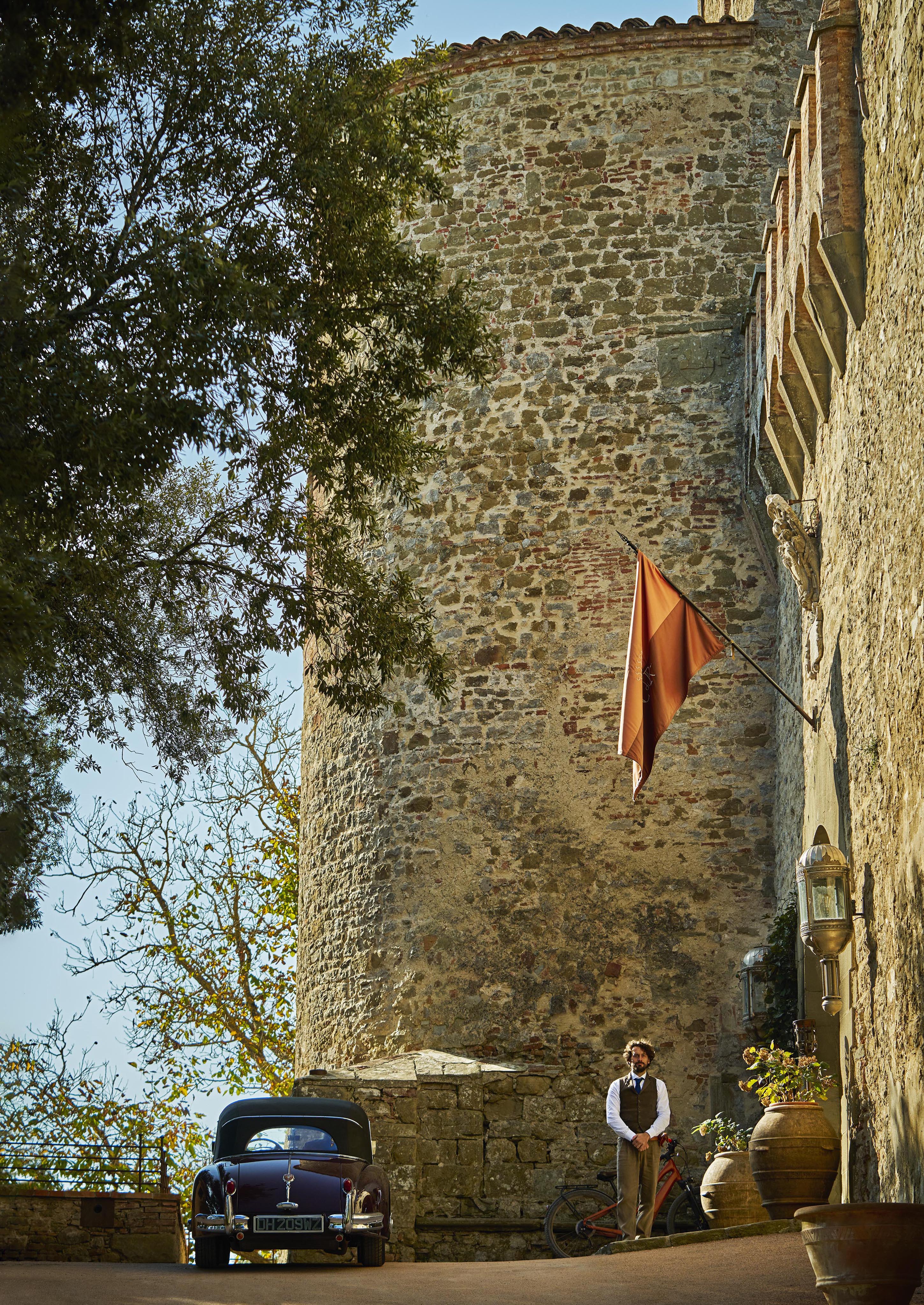 A man stands next to a vintage car and bicycle by a large stone building with a flag.