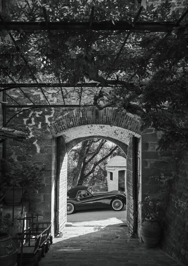 A vintage car is parked outside a brick archway, surrounded by stone walls and leafy vines overhead. Black and white image.