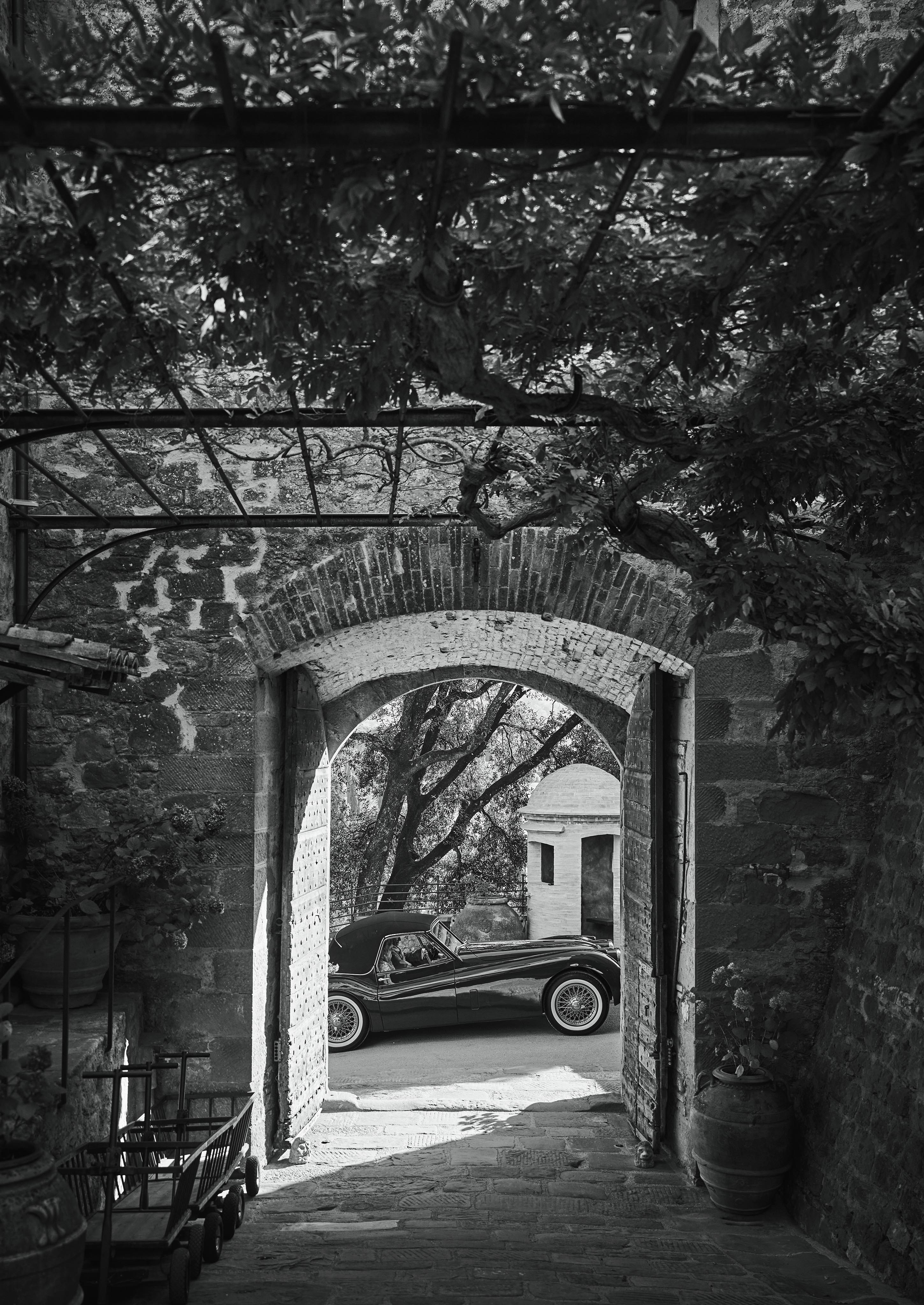 A vintage car is parked outside a brick archway, surrounded by stone walls and leafy vines overhead. Black and white image.