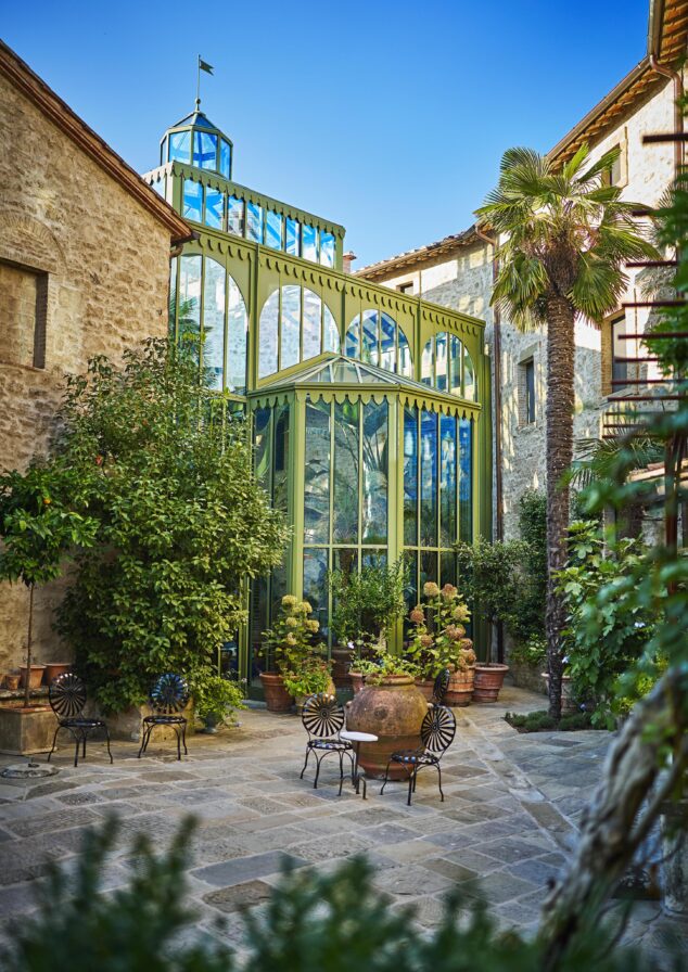 Stone courtyard with a green, glass-paneled structure surrounded by lush plants. Black wrought iron chairs and a round table are set in the center. Tall palm tree nearby. Clear blue sky overhead.