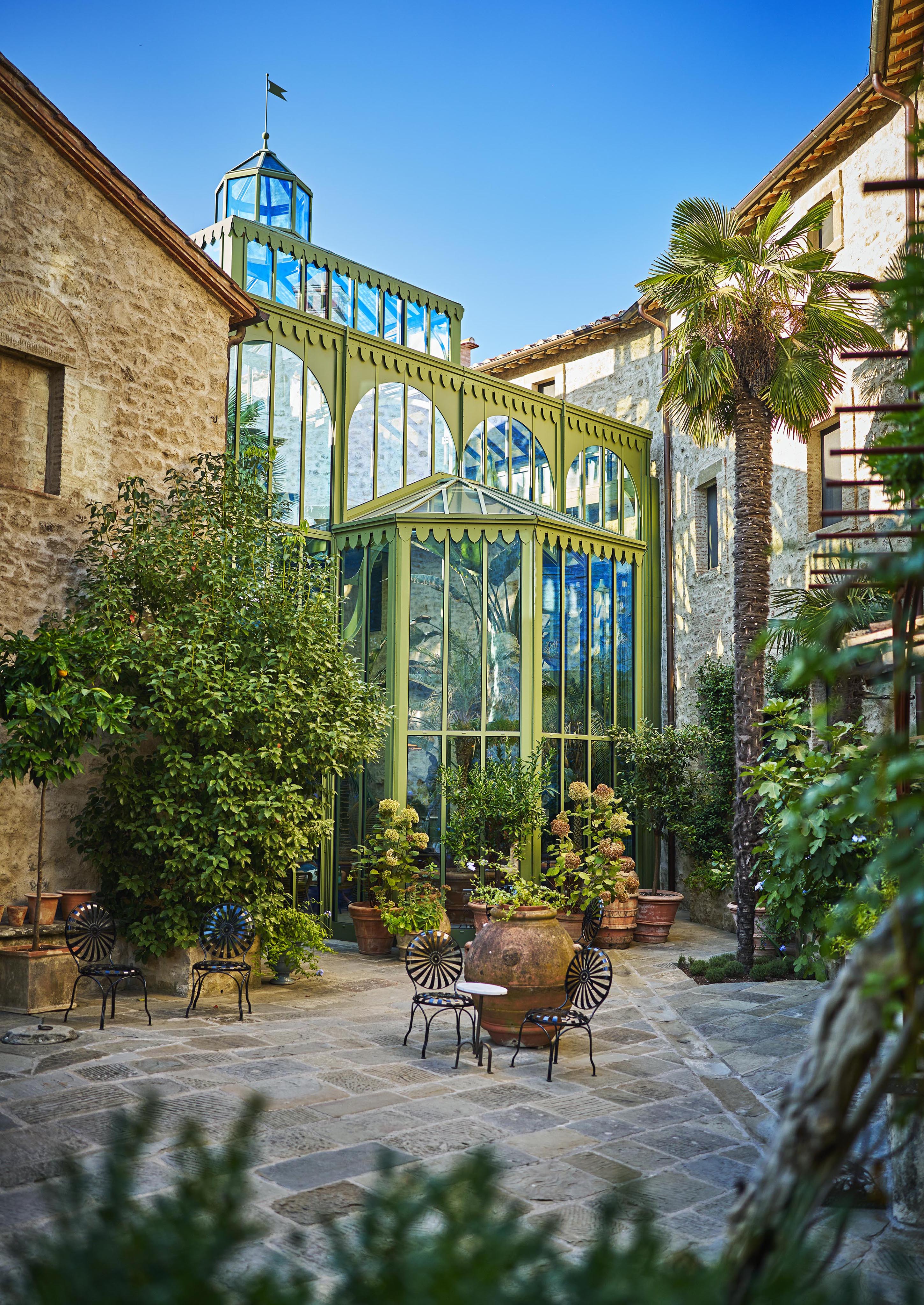 Stone courtyard with a green, glass-paneled structure surrounded by lush plants. Black wrought iron chairs and a round table are set in the center. Tall palm tree nearby. Clear blue sky overhead.
