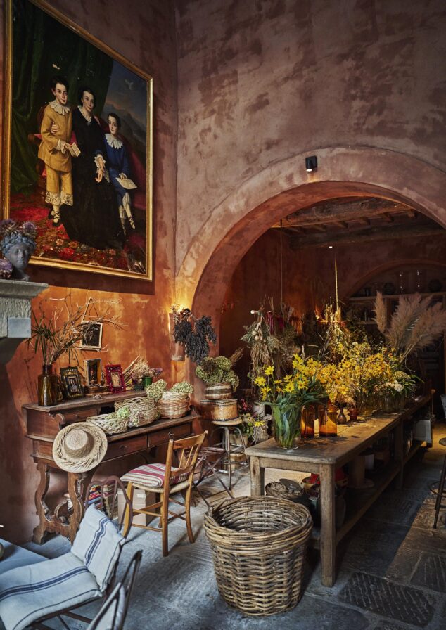 A rustic room with a large painting on the wall, a wooden table filled with flowers, chairs, a basket, and a side table with framed pictures. The walls are painted in a warm, earthy tone.