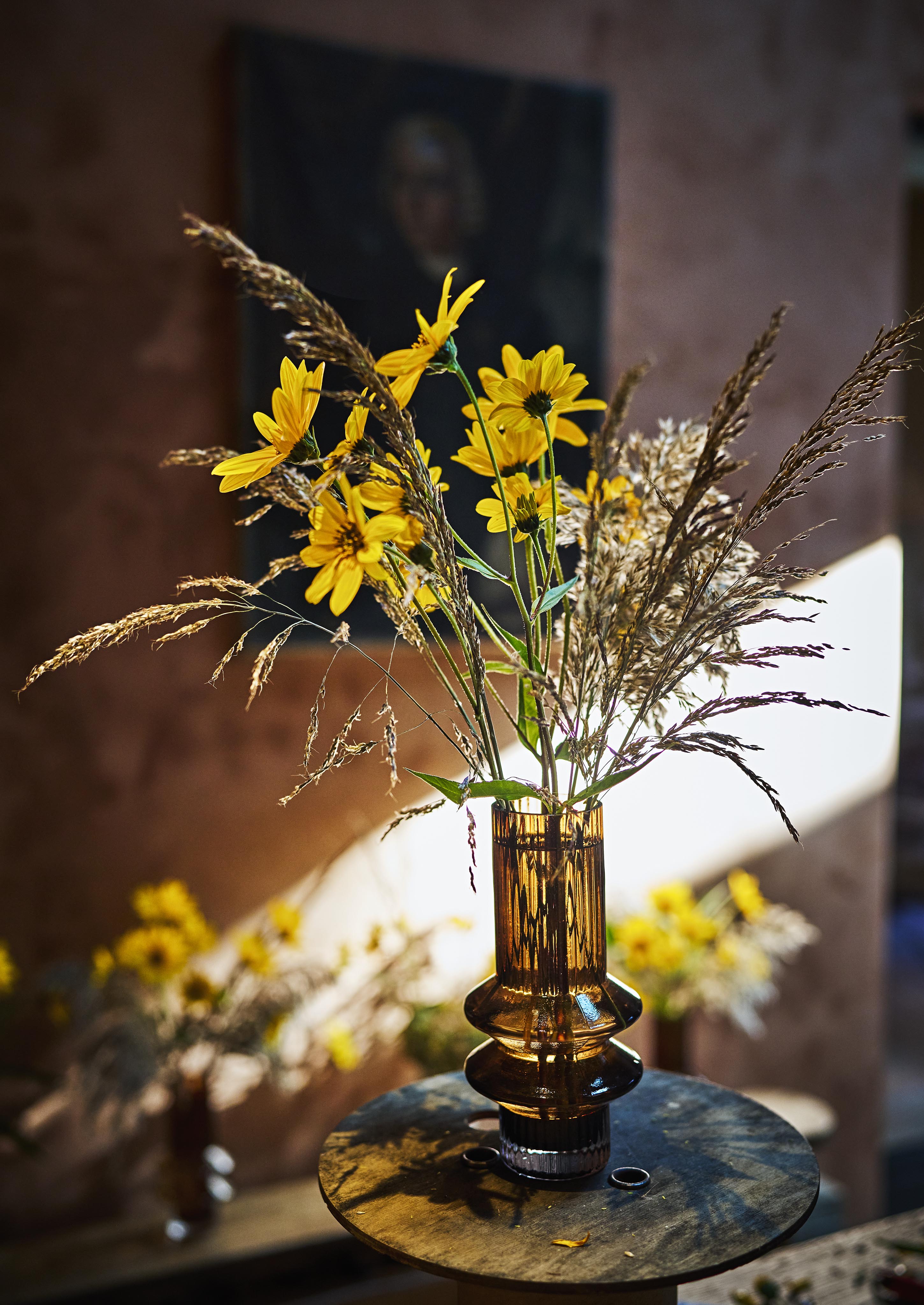 A brown glass vase holds yellow flowers and tall grass on a table. Sunlight illuminates the scene, casting soft shadows. A blurred background features additional vases and a dark painting.