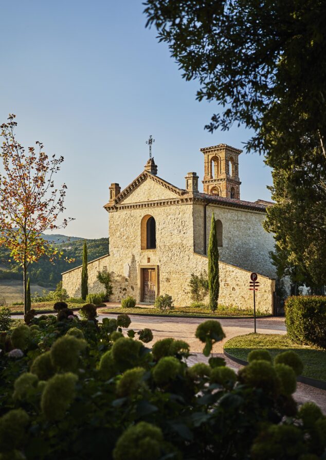 Old stone building with a bell tower and arched windows, surrounded by trees and greenery, under a clear blue sky.