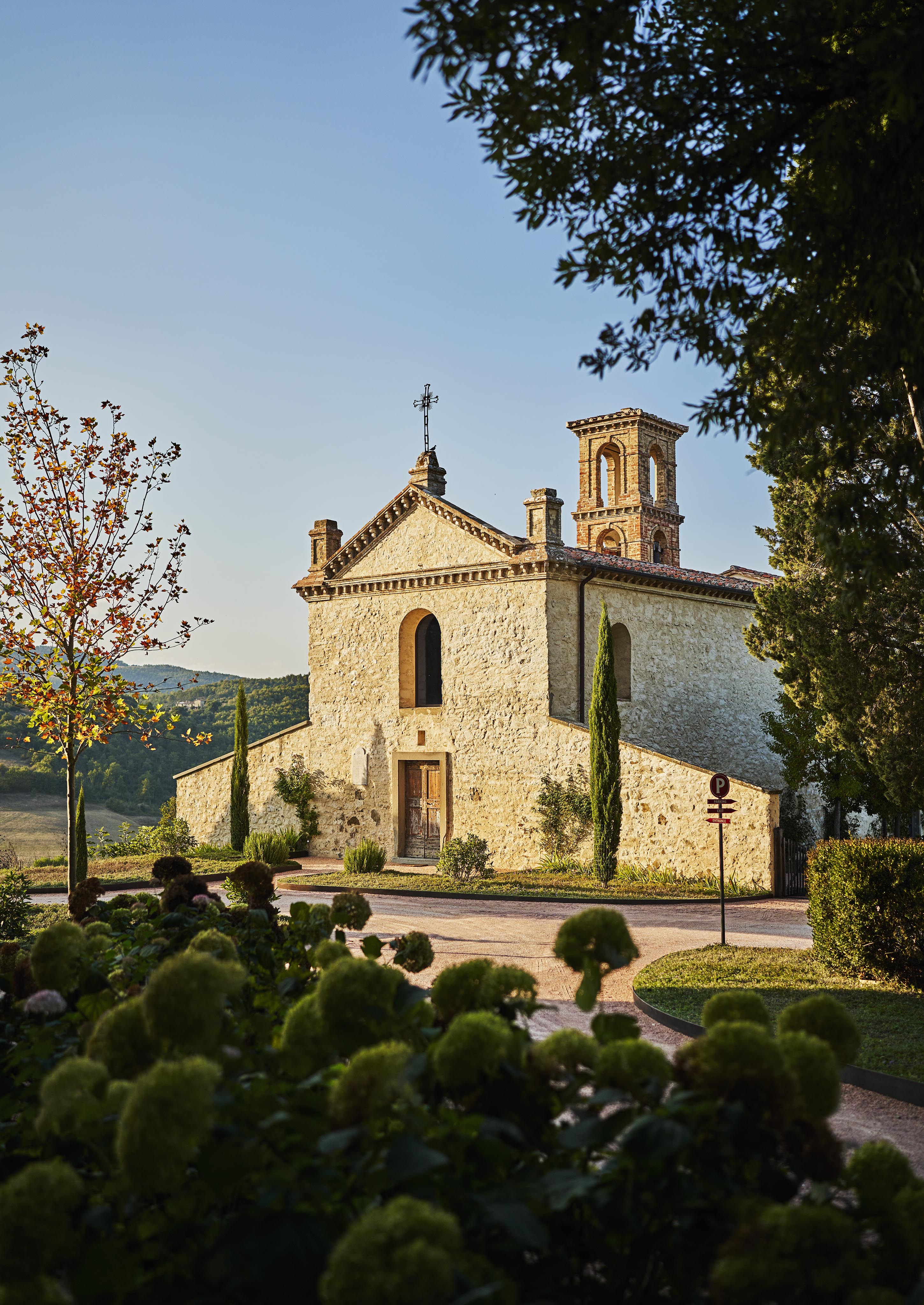 Old stone building with a bell tower and arched windows, surrounded by trees and greenery, under a clear blue sky.