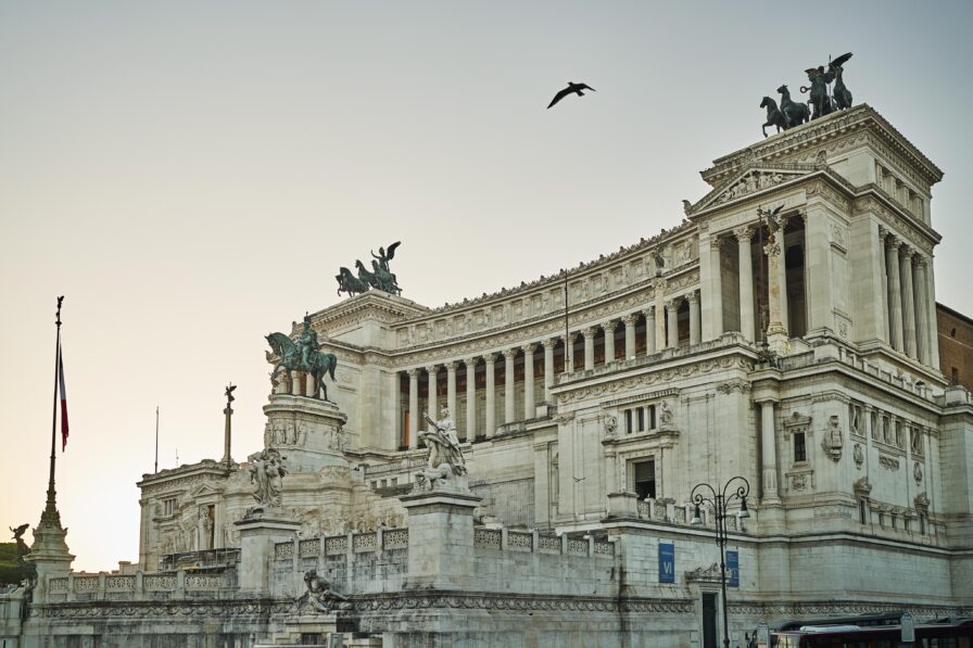 Large neoclassical building with columns and statues, featuring a silhouette of a bird flying overhead against a clear sky. A flag is visible to the left.