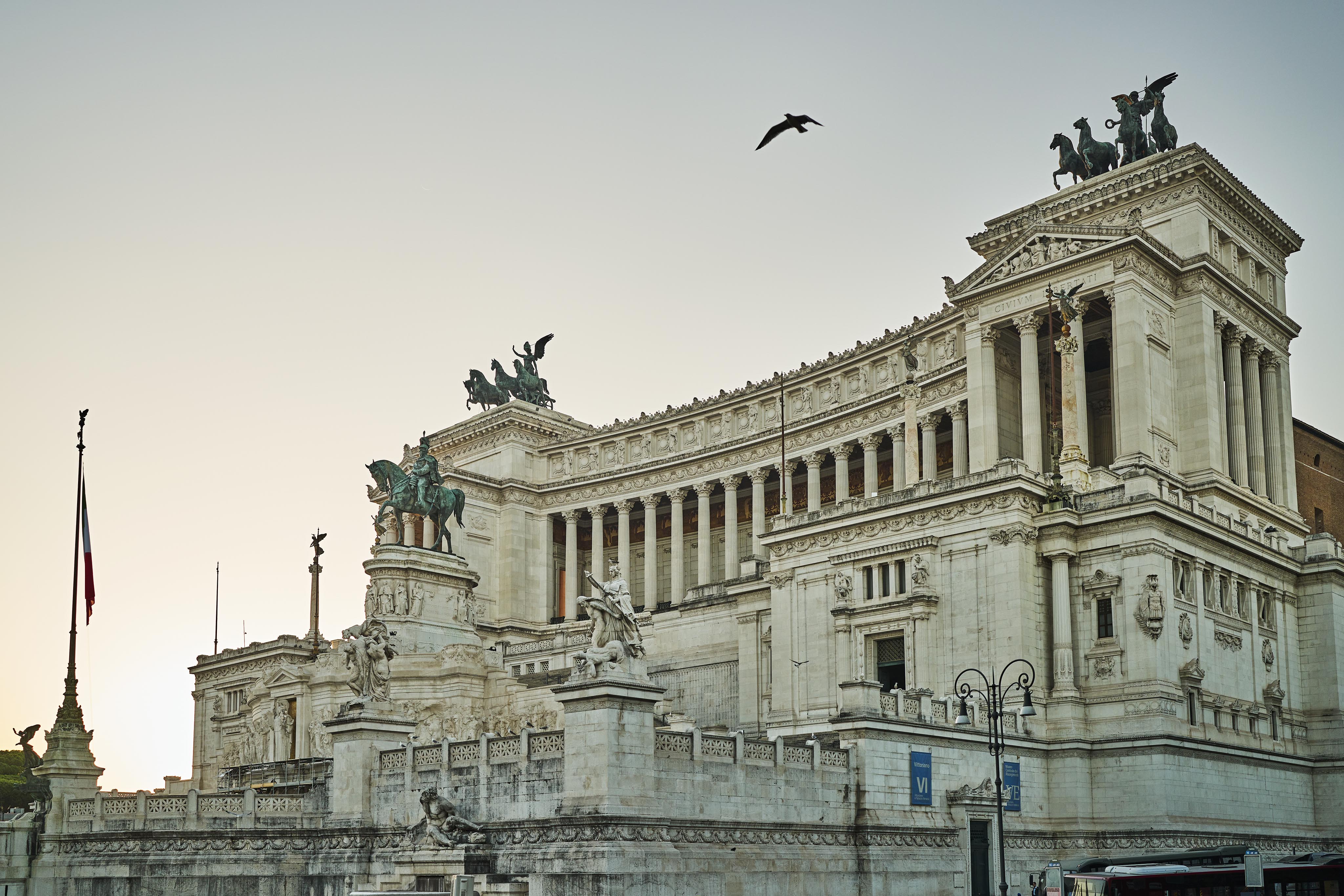 Large neoclassical building with columns and statues, featuring a silhouette of a bird flying overhead against a clear sky. A flag is visible to the left.