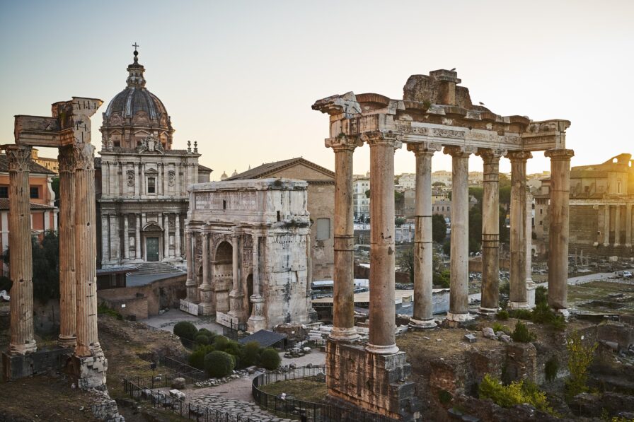 Ancient Roman ruins with tall columns and arches at sunset, beside a historic building with a dome.