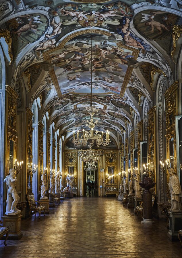 Ornate corridor with a high ceiling featuring detailed frescoes, chandeliers, sculptures along the walls, and a reflection on the polished floor.