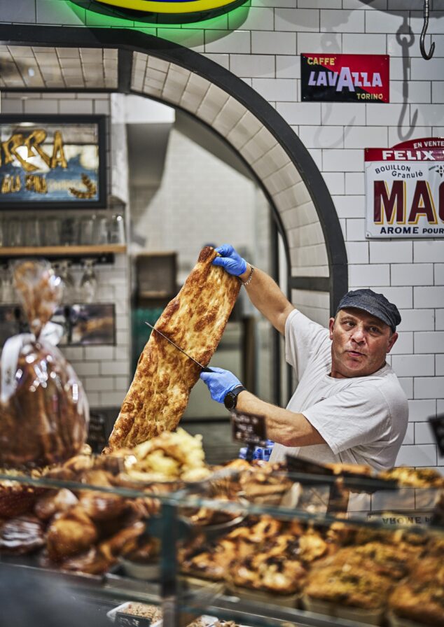 A baker in a white shirt and cap, wearing blue gloves, cuts a large piece of focaccia in a bakery with tiled walls and various baked goods on display.