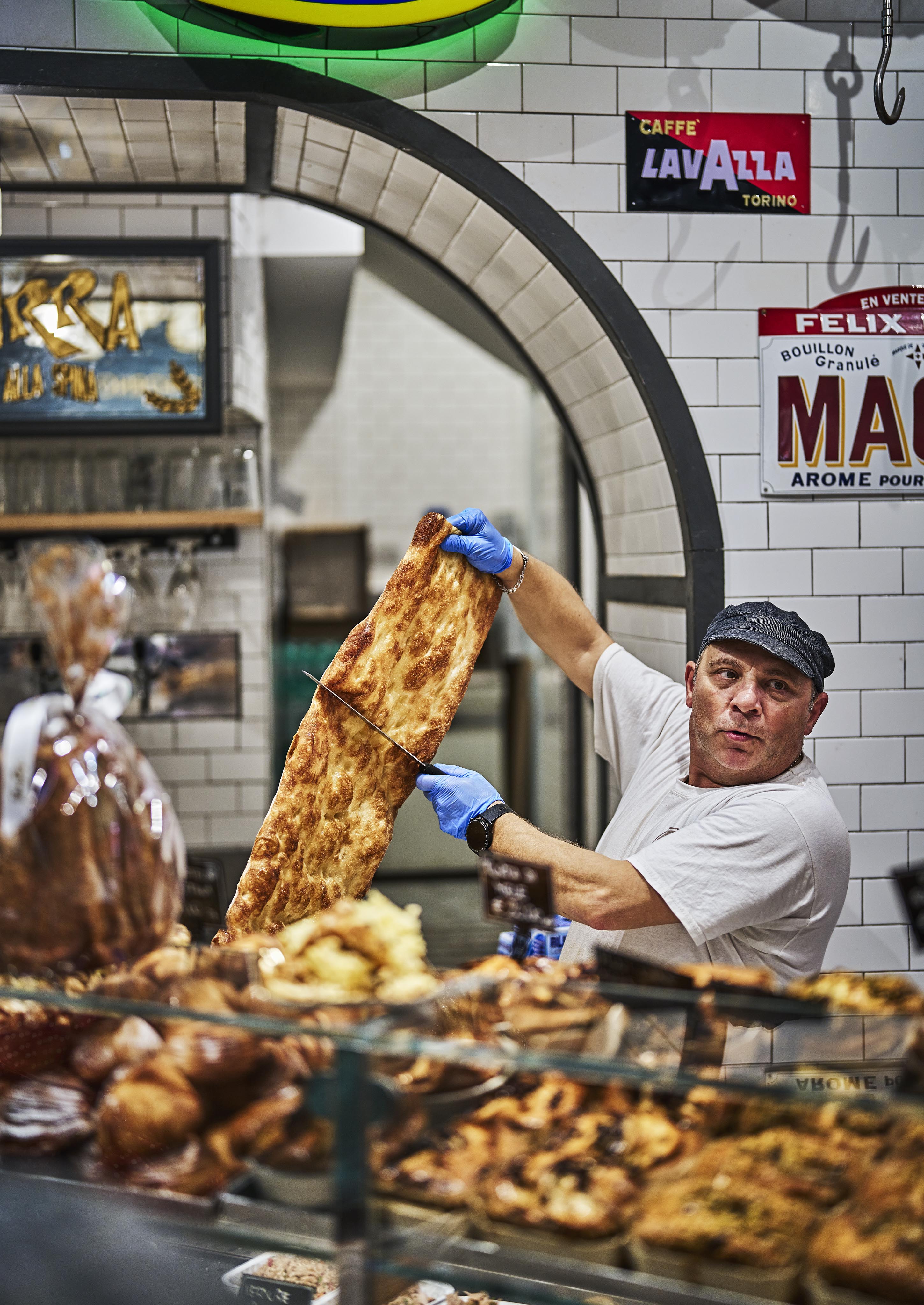 A baker in a white shirt and cap, wearing blue gloves, cuts a large piece of focaccia in a bakery with tiled walls and various baked goods on display.