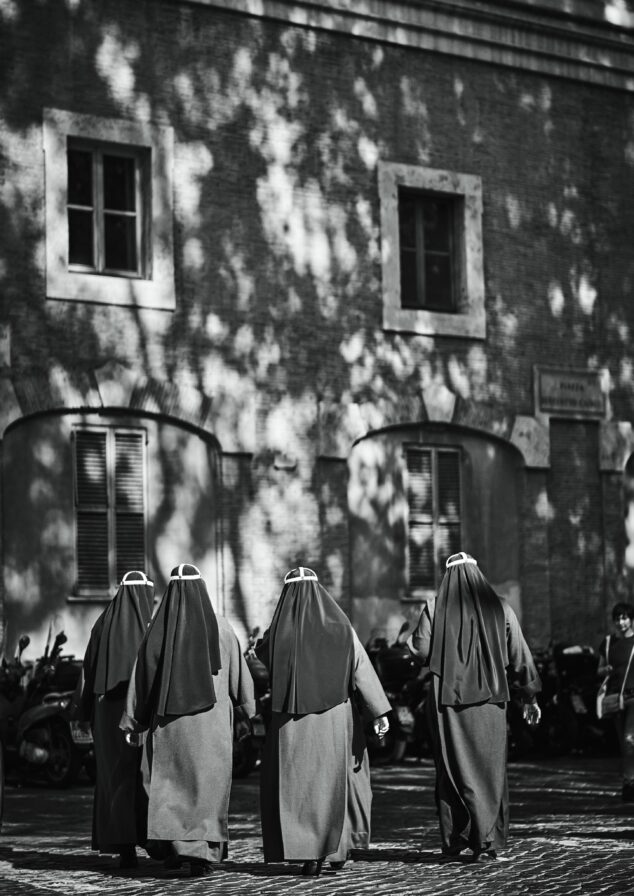 Four nuns walk side by side on a cobblestone street in front of a brick building with windows. Their shadows are visible on the ground. A line of parked scooters is in the background.