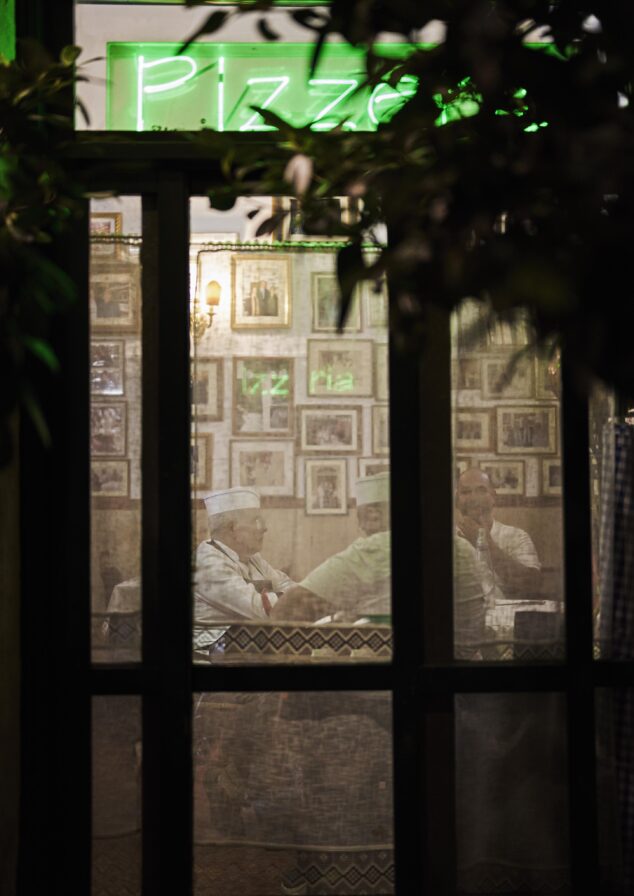 Several people sit around a table in a dimly lit room, visible through a window. A neon sign above reads "Pizza." Walls are decorated with framed photos.