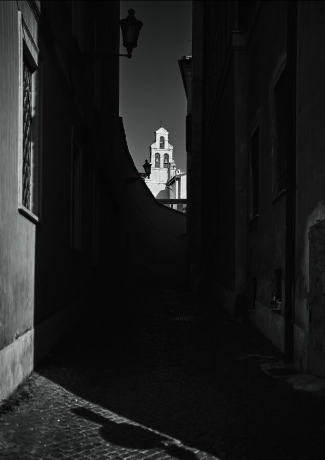 A narrow, dark alley frames a brightly lit bell tower in the background. Cobblestone ground and building shadows dominate the foreground.