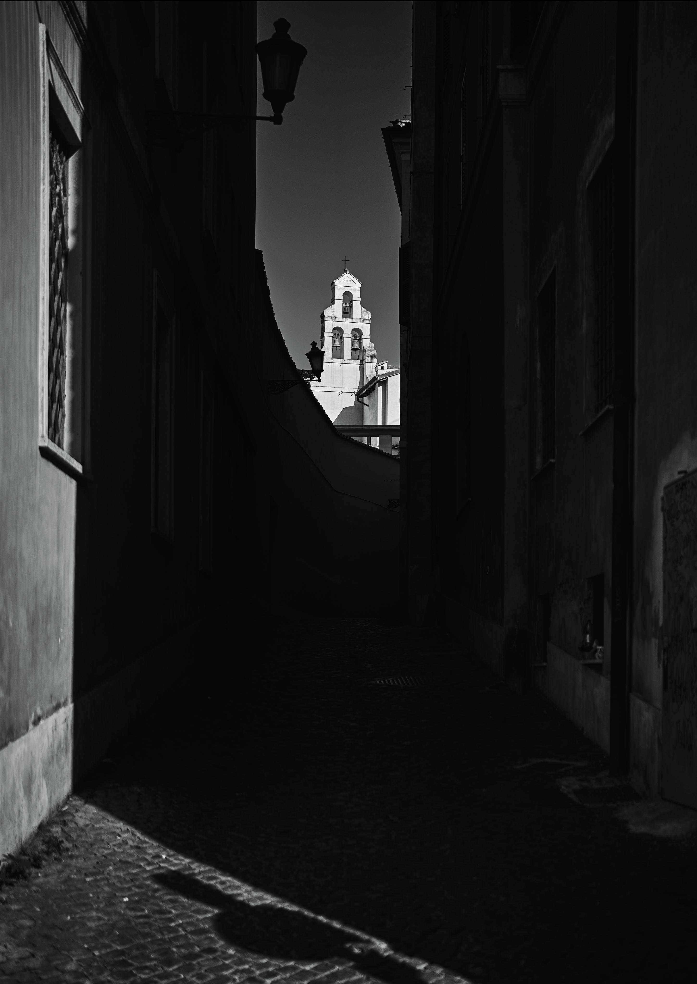 A narrow, dark alley frames a brightly lit bell tower in the background. Cobblestone ground and building shadows dominate the foreground.