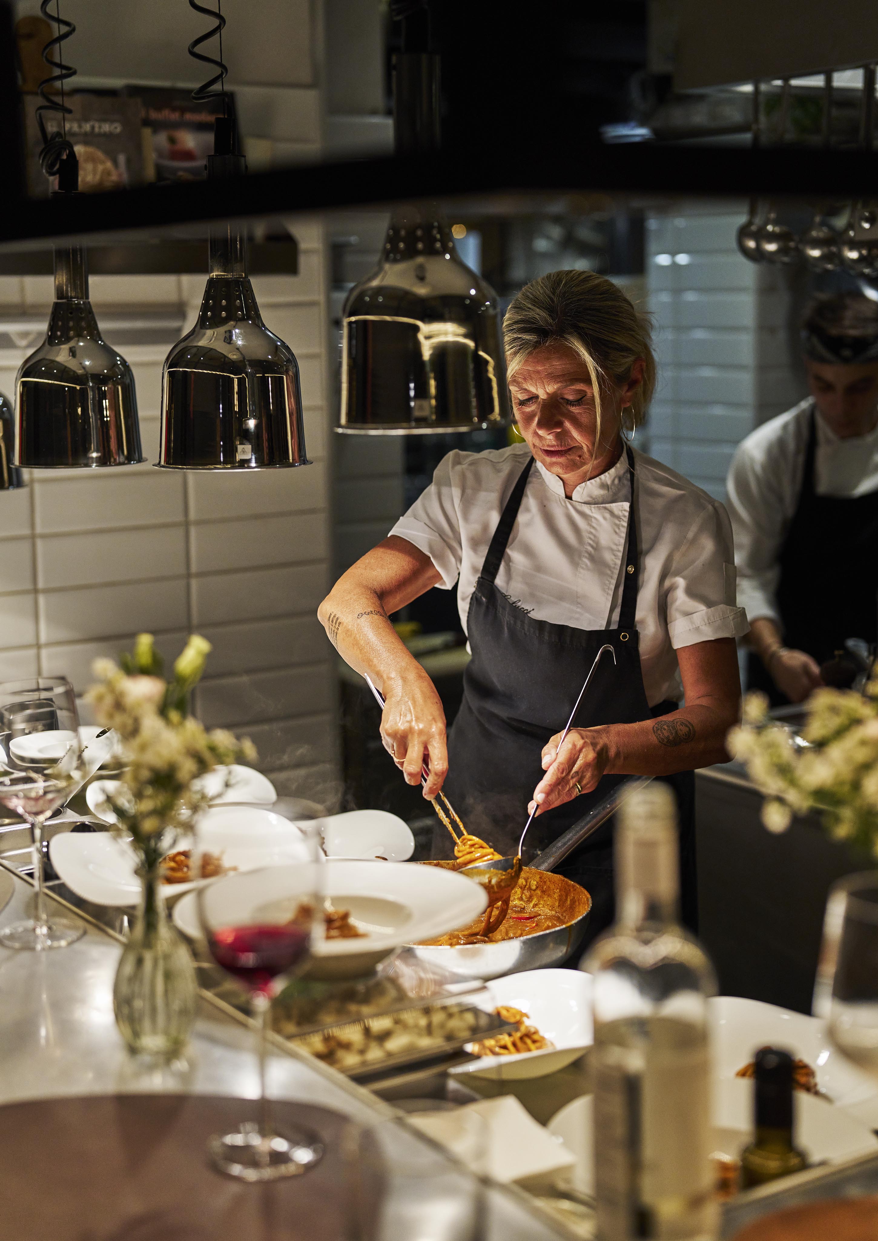 A chef in a white shirt and dark apron skillfully plates food at a restaurant kitchen counter, surrounded by dishes and wine glasses, with dim lighting above.