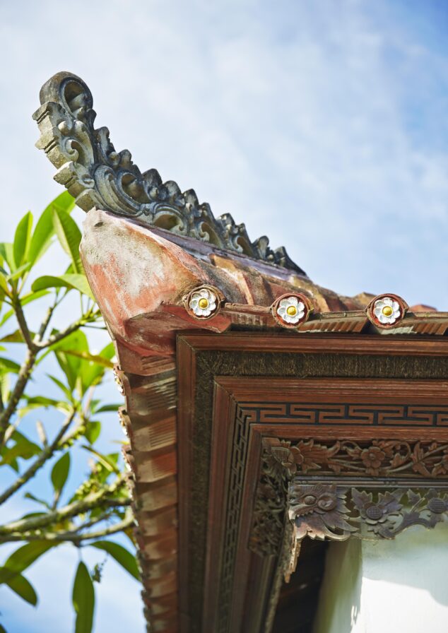 Close-up of a traditional Asian roof corner with ornate details, under a blue sky, and a leafy branch in the background.