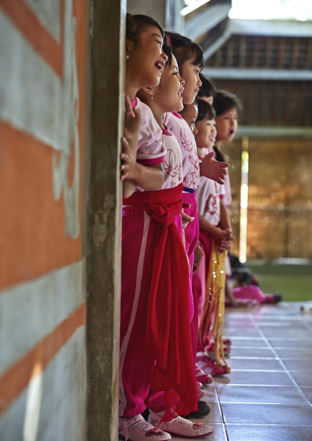 Children in pink outfits stand in a line against a wall, smiling and watching something to their right.