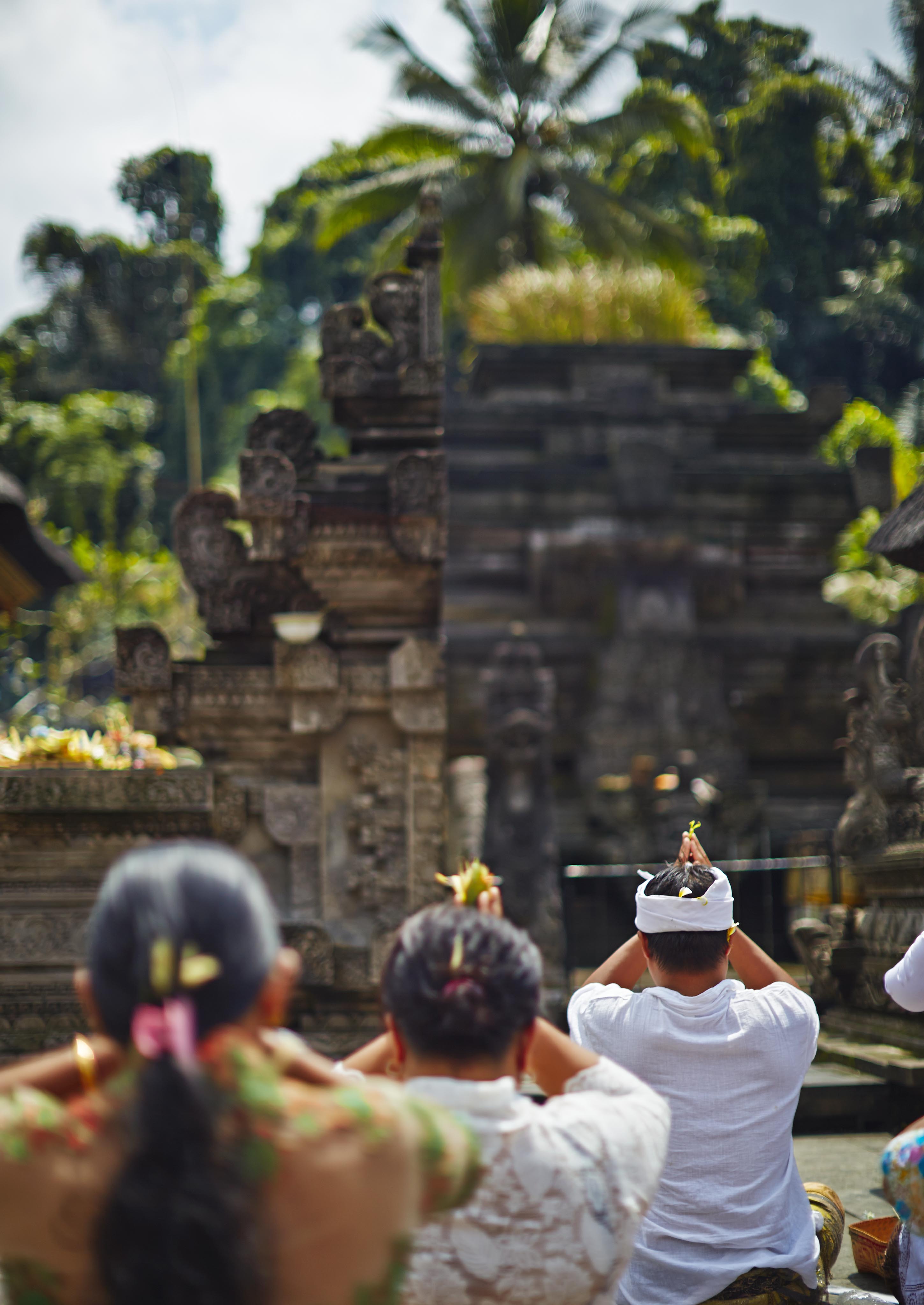 Group of people in traditional attire facing a Balinese temple, hands raised in prayer. Lush greenery and stone carvings visible in the background.