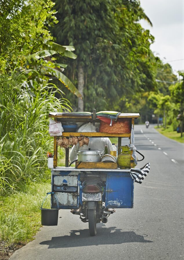 Food cart on a motorcycle driving down a rural road lined with greenery, carrying various cooking items and supplies.