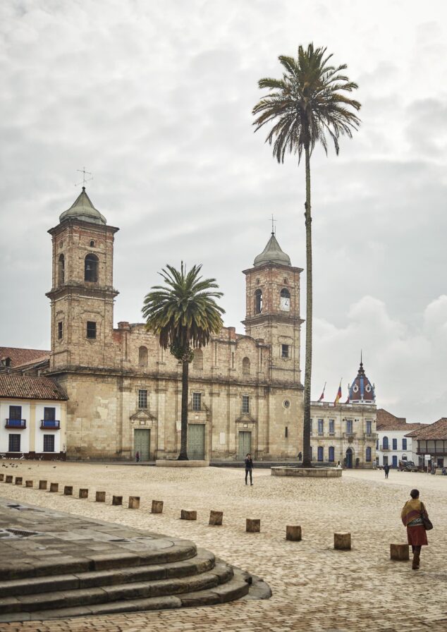 A historic church with two bell towers stands gracefully in a cobblestone plaza, flanked by tall palm trees reminiscent of South America's charm. A few people stroll nearby, while a building with a clock tower is visible in the background.