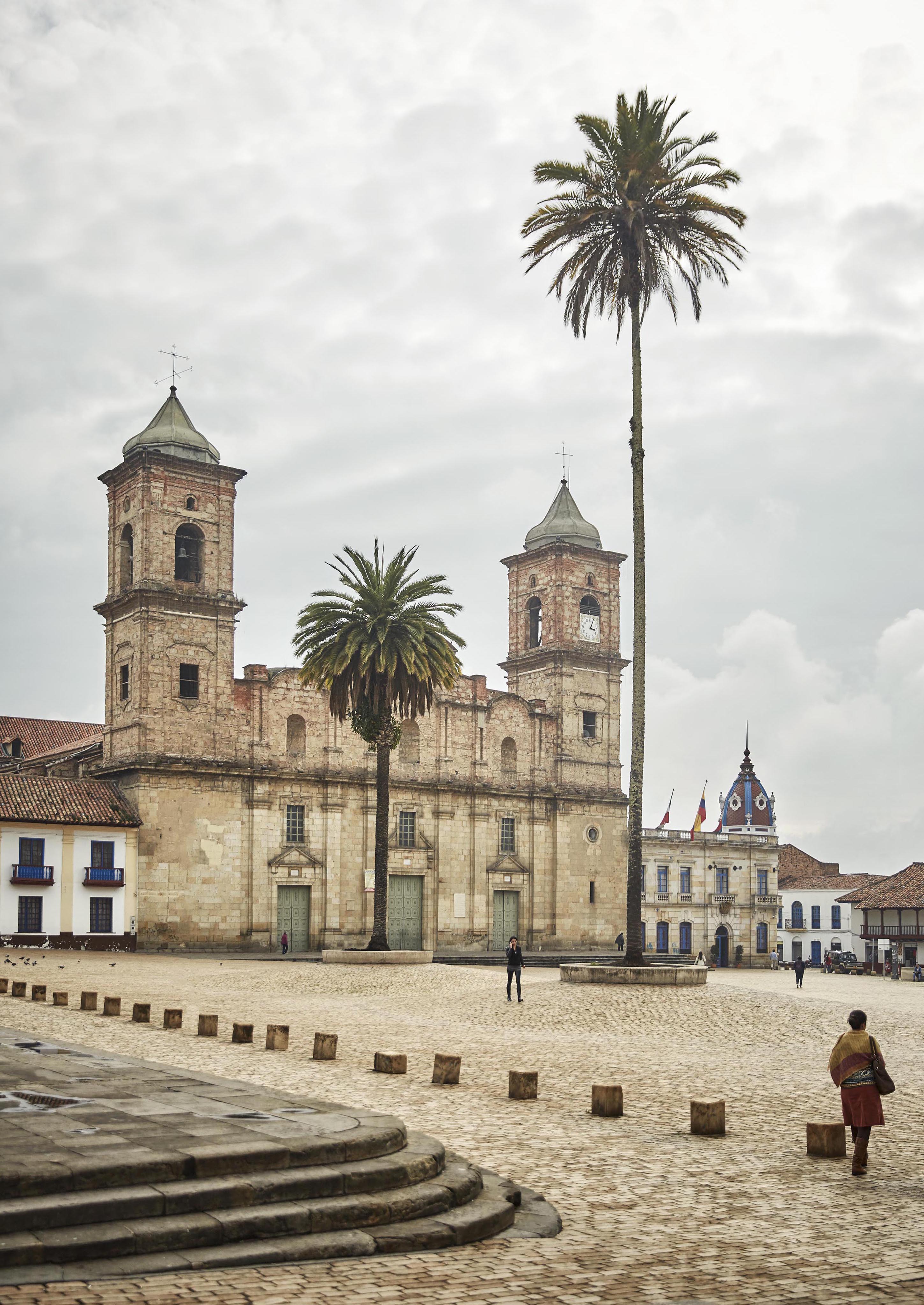 A historic church with two bell towers stands gracefully in a cobblestone plaza, flanked by tall palm trees reminiscent of South America's charm. A few people stroll nearby, while a building with a clock tower is visible in the background.