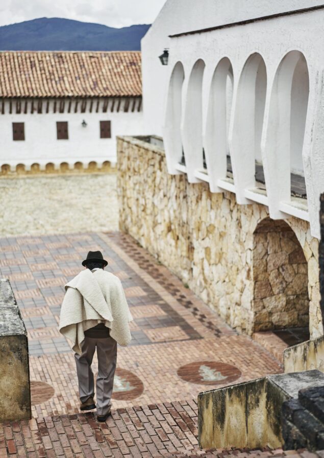 A person in a poncho and hat strolls down brick steps beside a white arched building with a tiled roof, evoking the charming architecture reminiscent of South America.