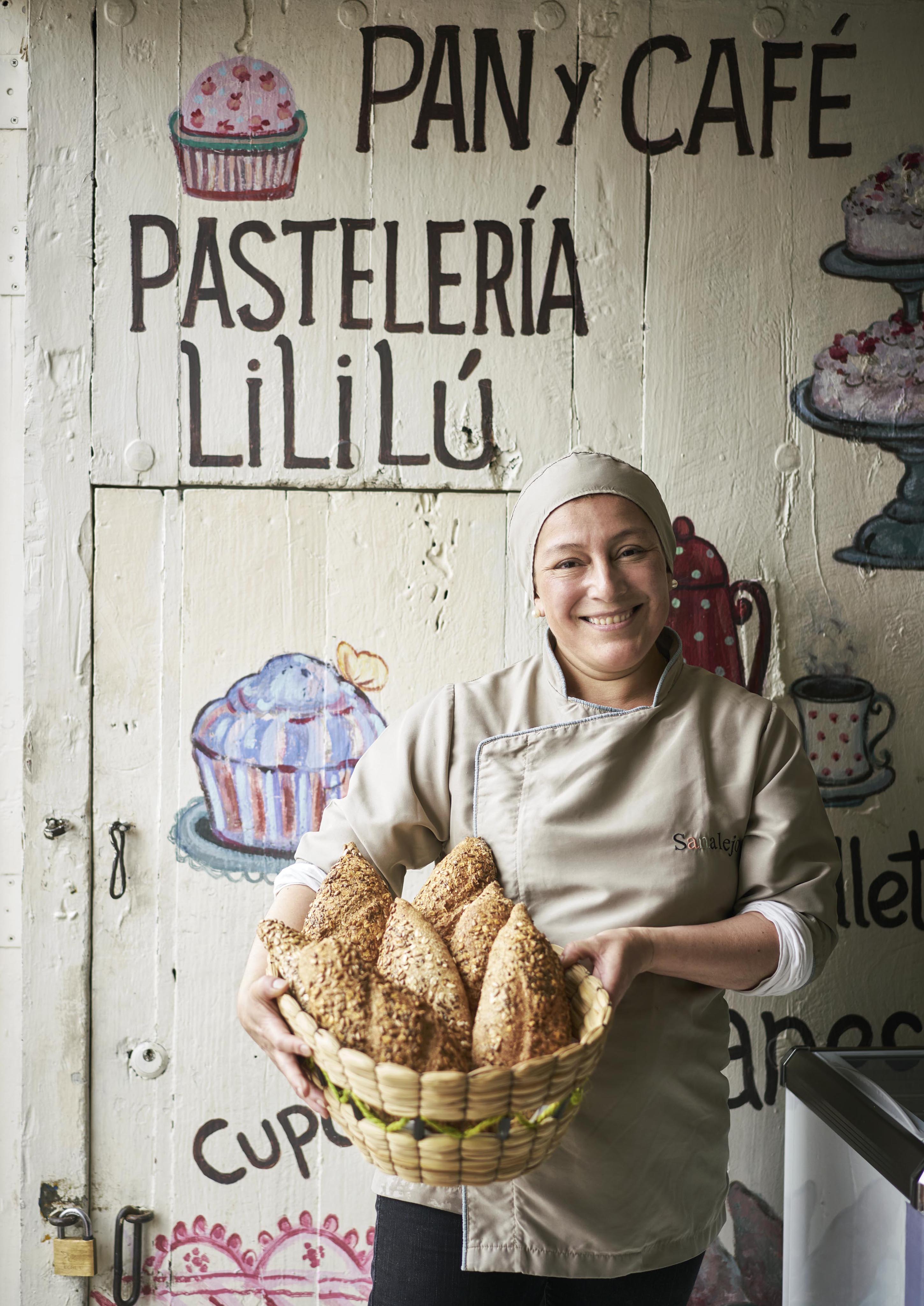 A smiling person in a chef's uniform holds a basket of bread in front of a bakery wall painted with pastries and the words "Pastelería Lili Lú" and "Pan y Café," capturing the vibrant flavors of Central America.
