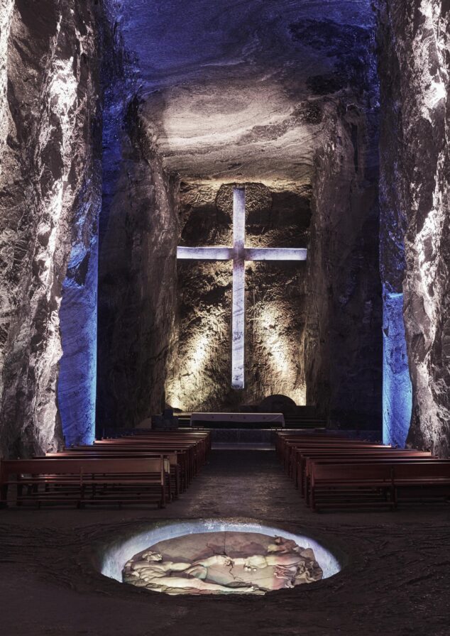 A large illuminated cross is mounted on a stone wall in an underground cathedral, reminiscent of the grand religious sites in South America. Rows of wooden pews face the altar, and the space is softly lit with blue and yellow lights.