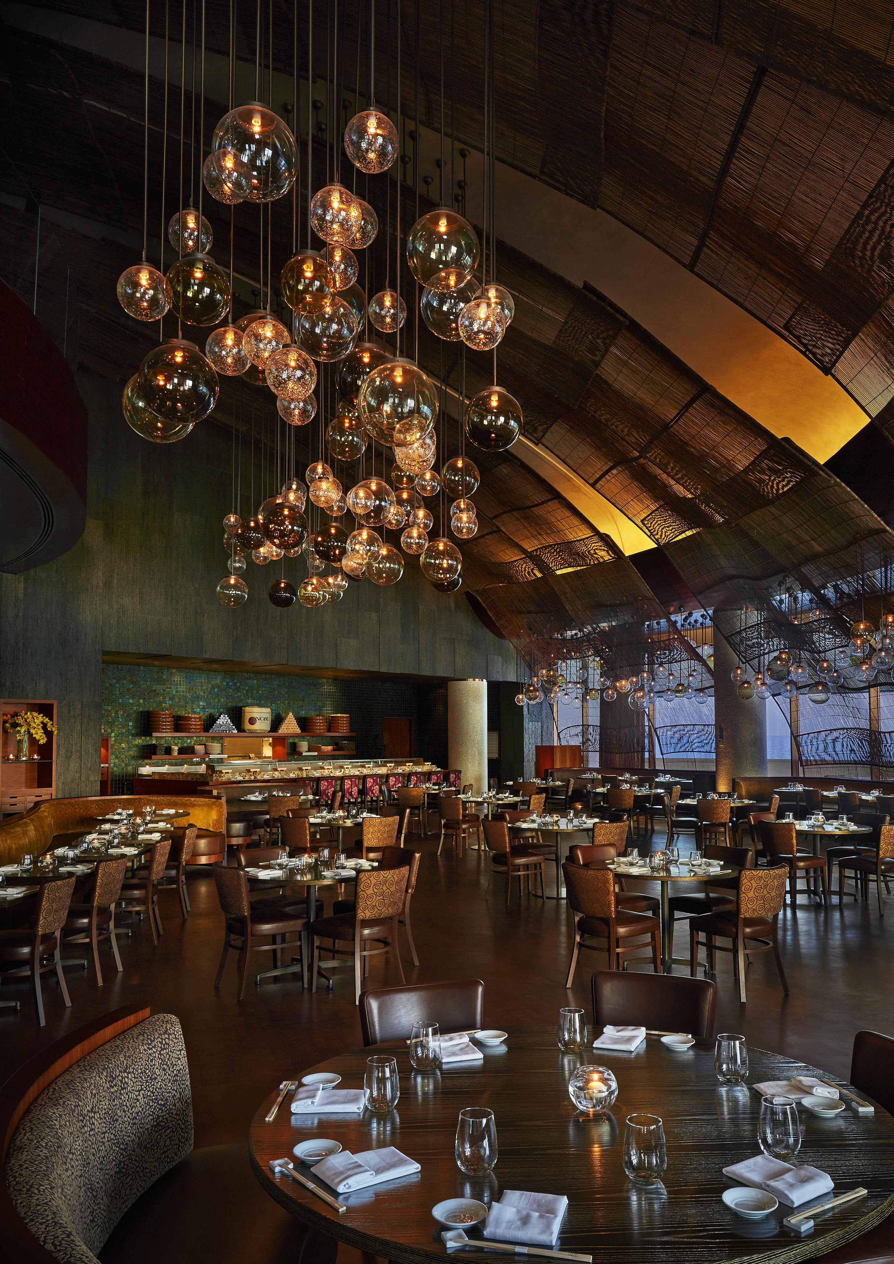The interior of Nobu located near Four Seasons DOHA, featuring a spacious, modern and warmly lit restaurant with wooden tables, neatly set place settings, and clusters of round clear glass pendant lights hanging from a high, arched ceiling.