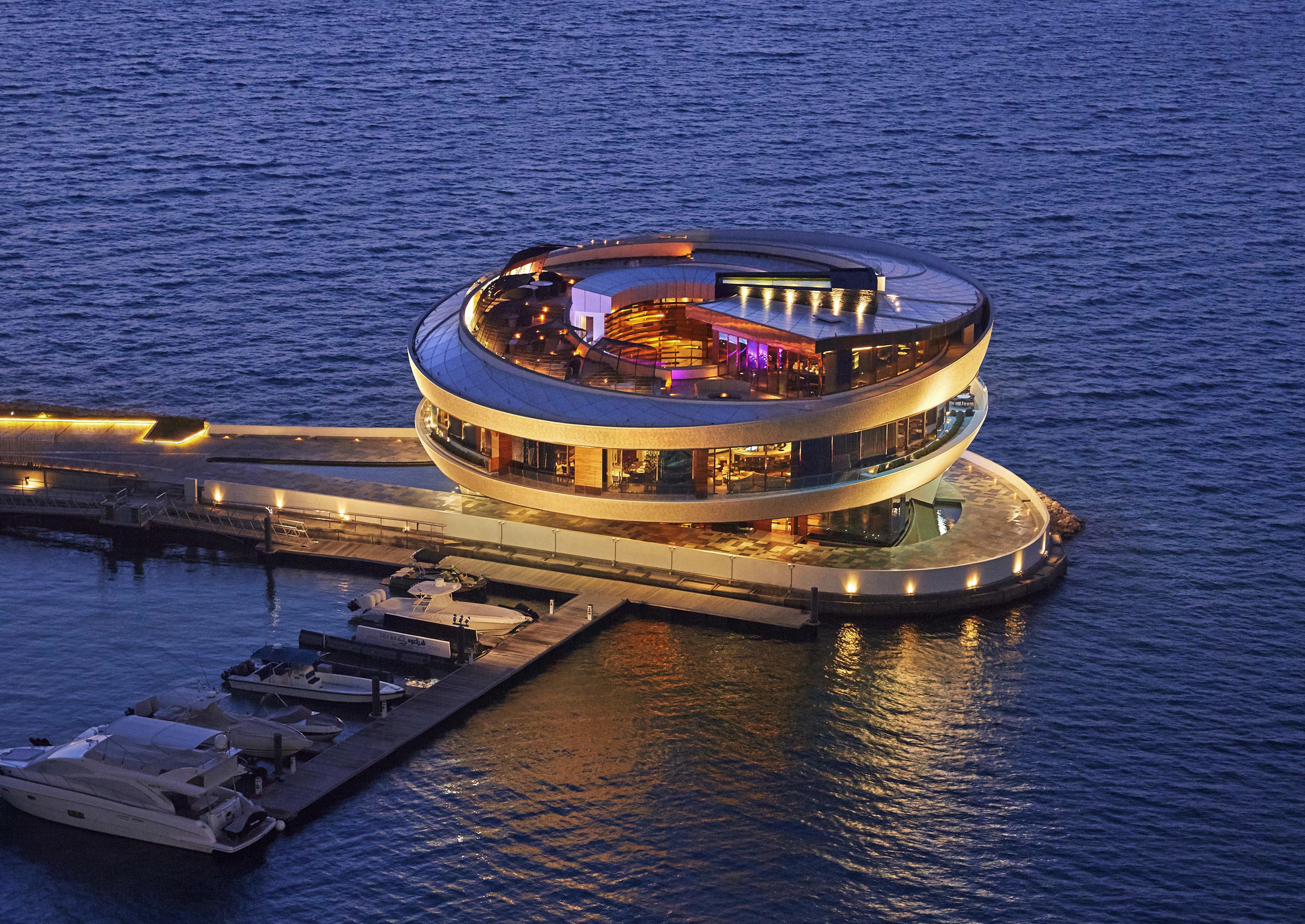 An aerial view of the Nobu restaurant within Four Seasons DOHA featuring a modern, circular spiraling structure with large windows and illuminated interiors sitting on a pier over the water, surrounded by boats and evening lights.