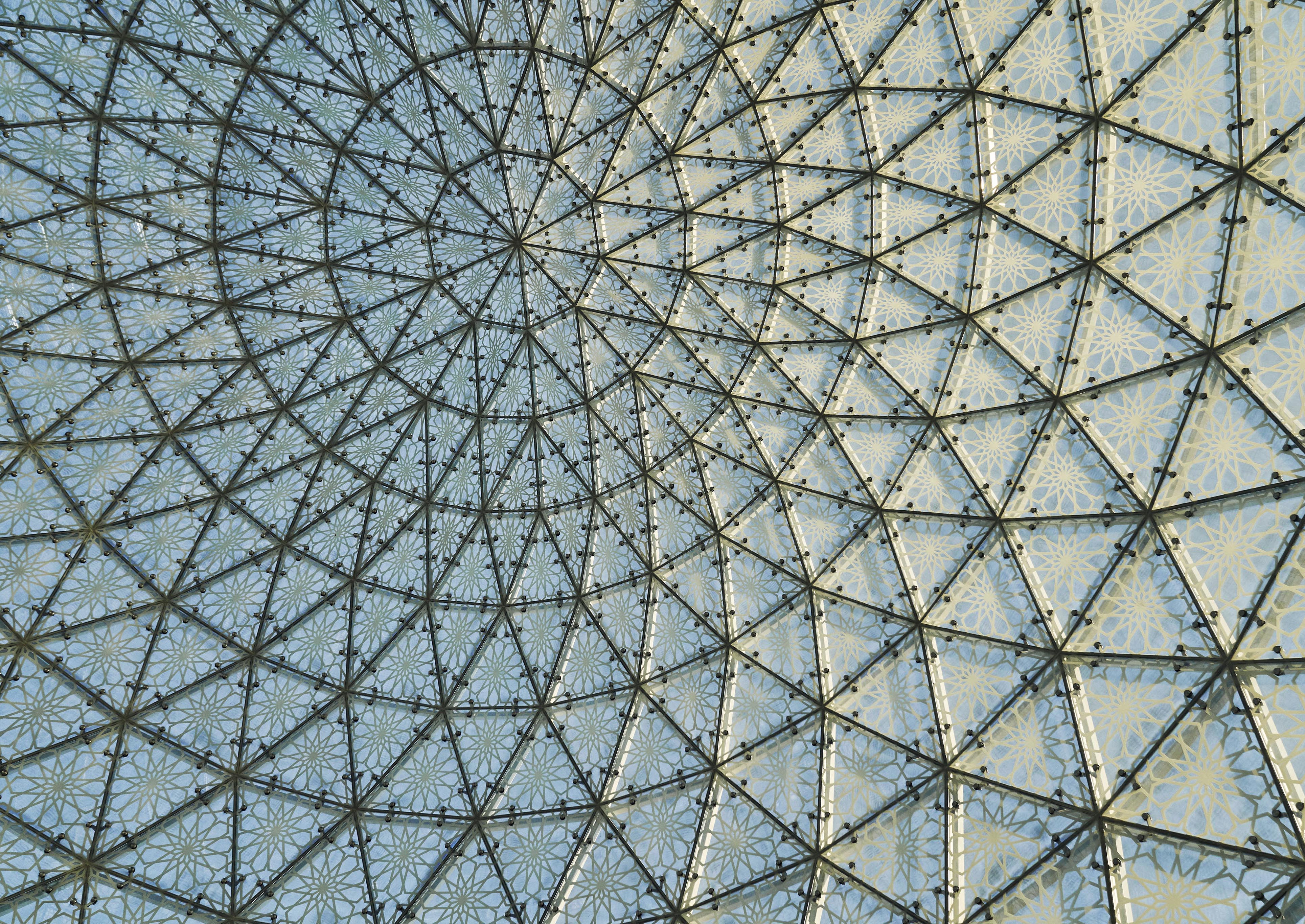 A detail view of landmark structure Abu Dhabi Grand Mosque with glass dome ceiling with a geometric metal framework and patterned panels forming a symmetrical, intricate design.