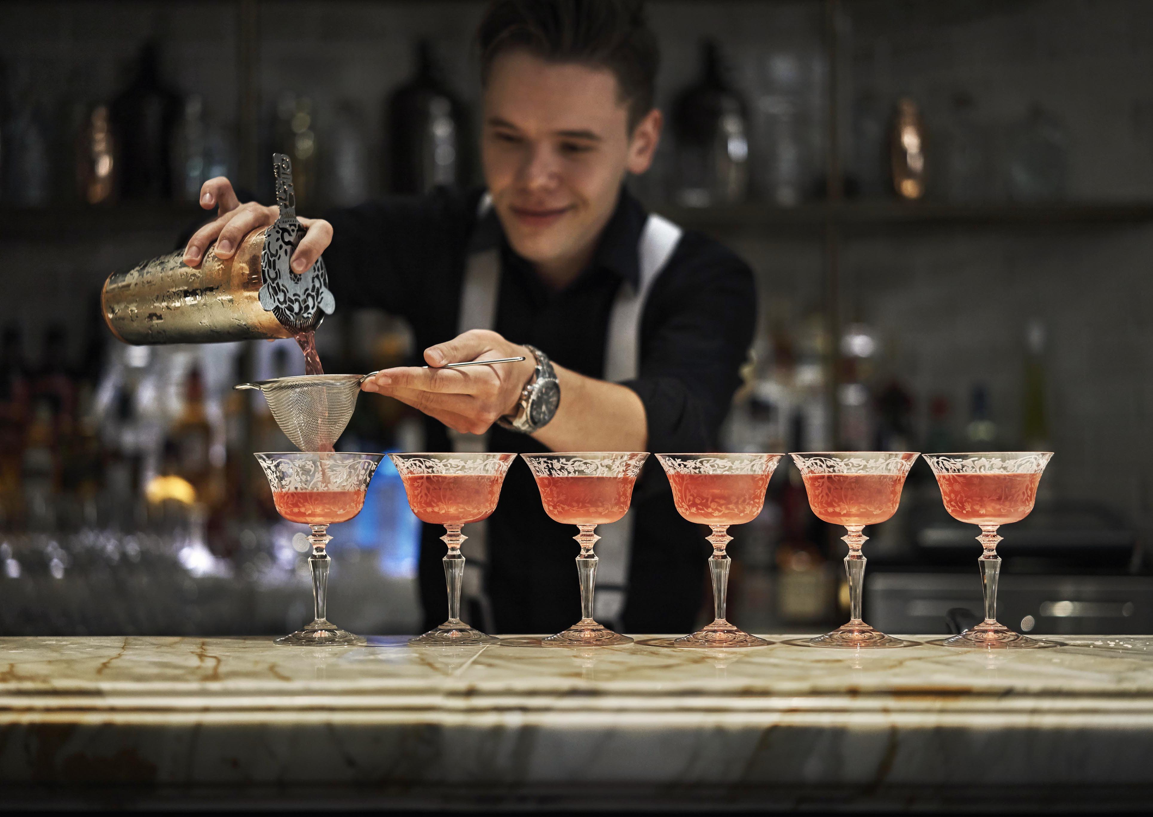 A delightful Abu Dhabi bar service staff pours elevated pink cocktails through a strainer into six coupe glasses lined up on a pristine marble bar counter.