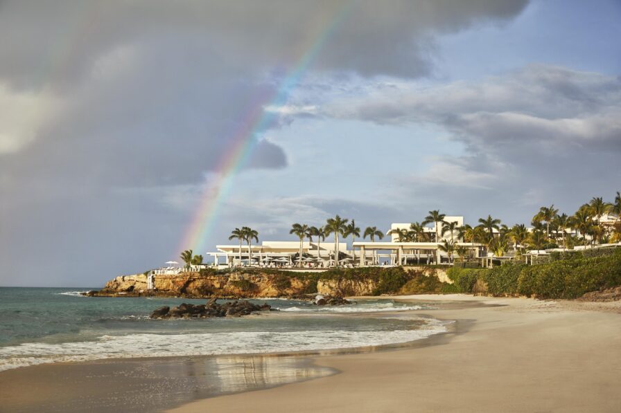 A scenic Anguilla beach resort exterior view with gentle waves, a rainbow in the cloudy sky, and a row of buildings and palm trees on a cliff.