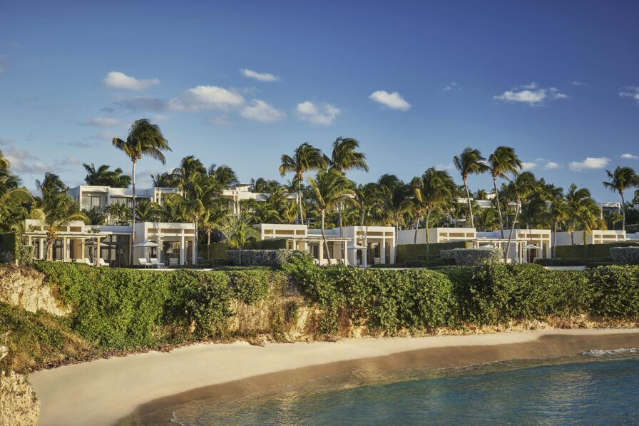 Beachfront Four Seasons Anguilla luxury resort villas surrounded by palm trees under a clear blue sky, with sandy beach and ocean in the foreground.