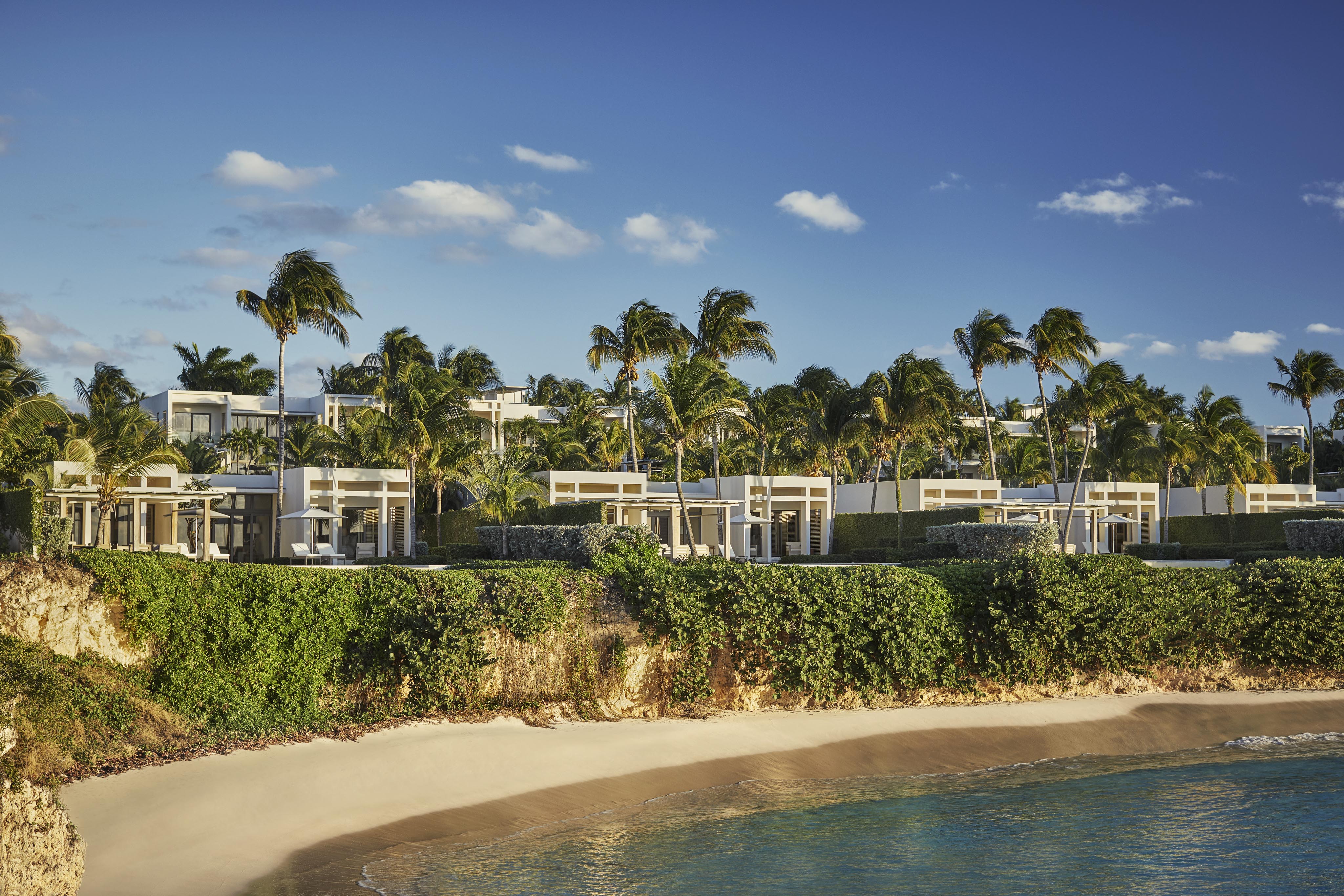 Beachfront Four Seasons Anguilla luxury resort villas surrounded by palm trees under a clear blue sky, with sandy beach and ocean in the foreground.