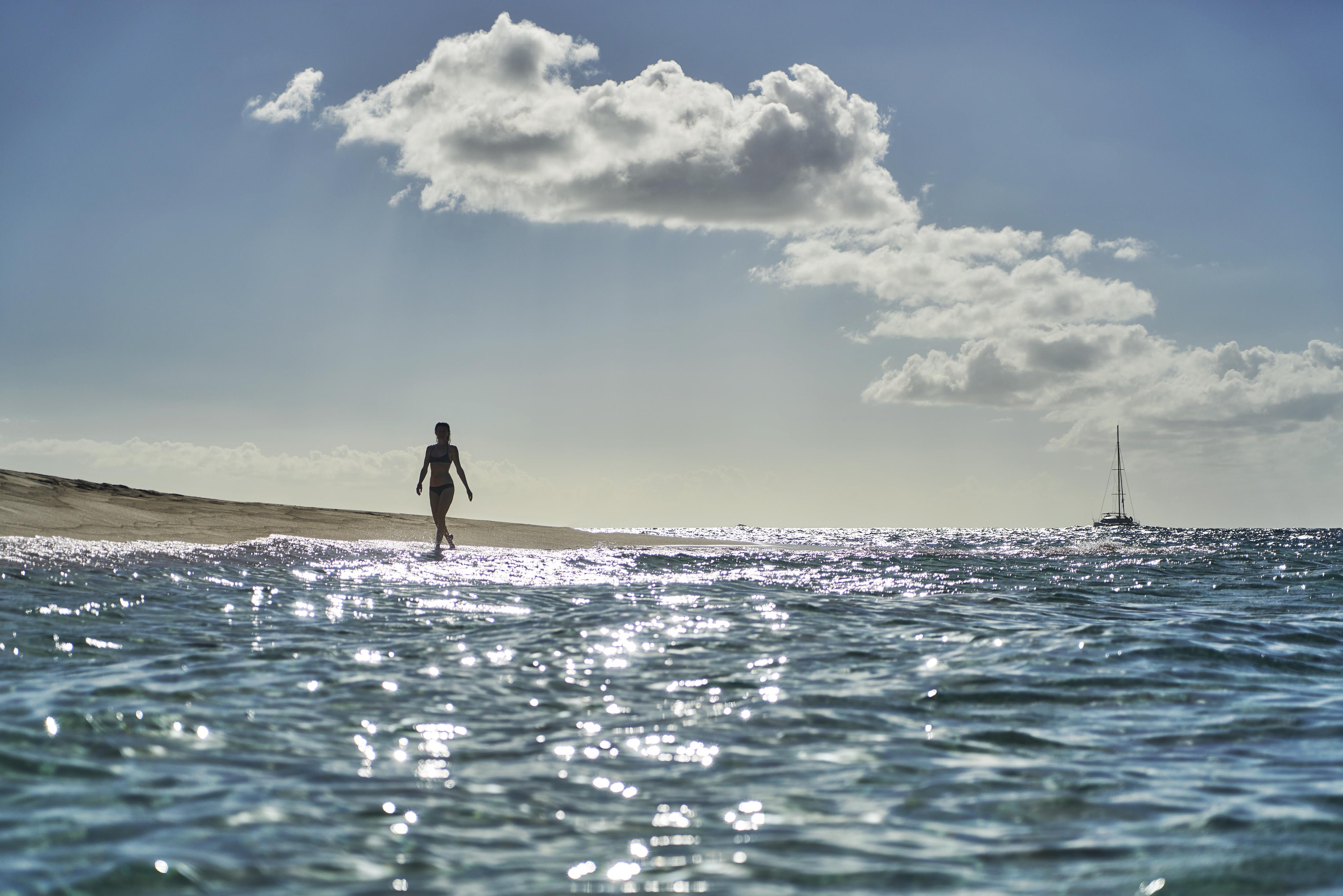 Person walks along a sunlit beach shoreline with a sailboat in the distance under a partly cloudy sky.