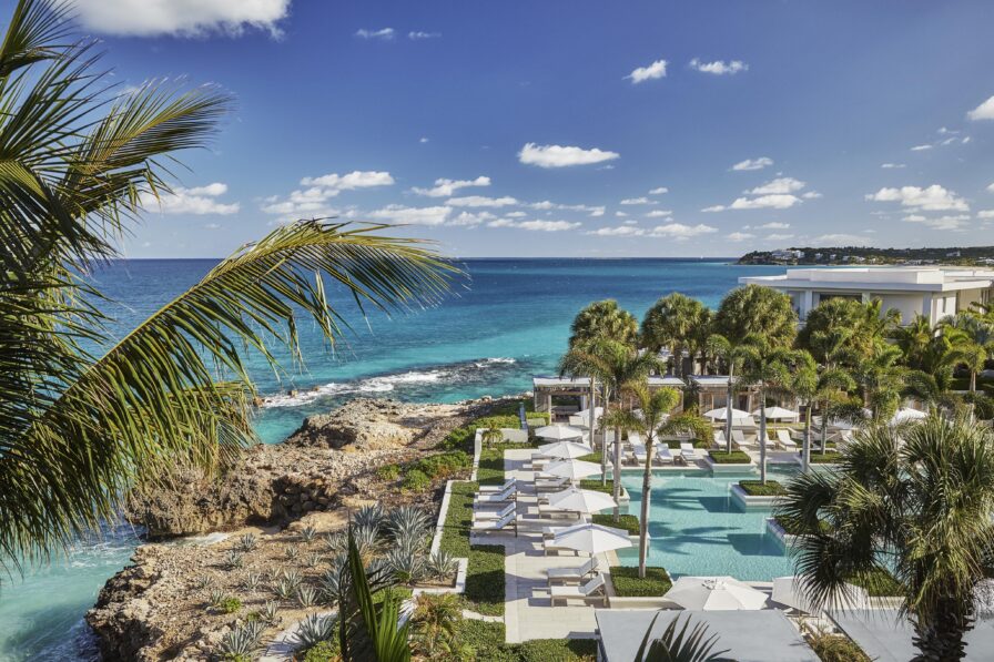 Anguilla Four Seasons resort pool area with lounge chairs and umbrellas overlooks turquoise ocean water under a clear blue sky.