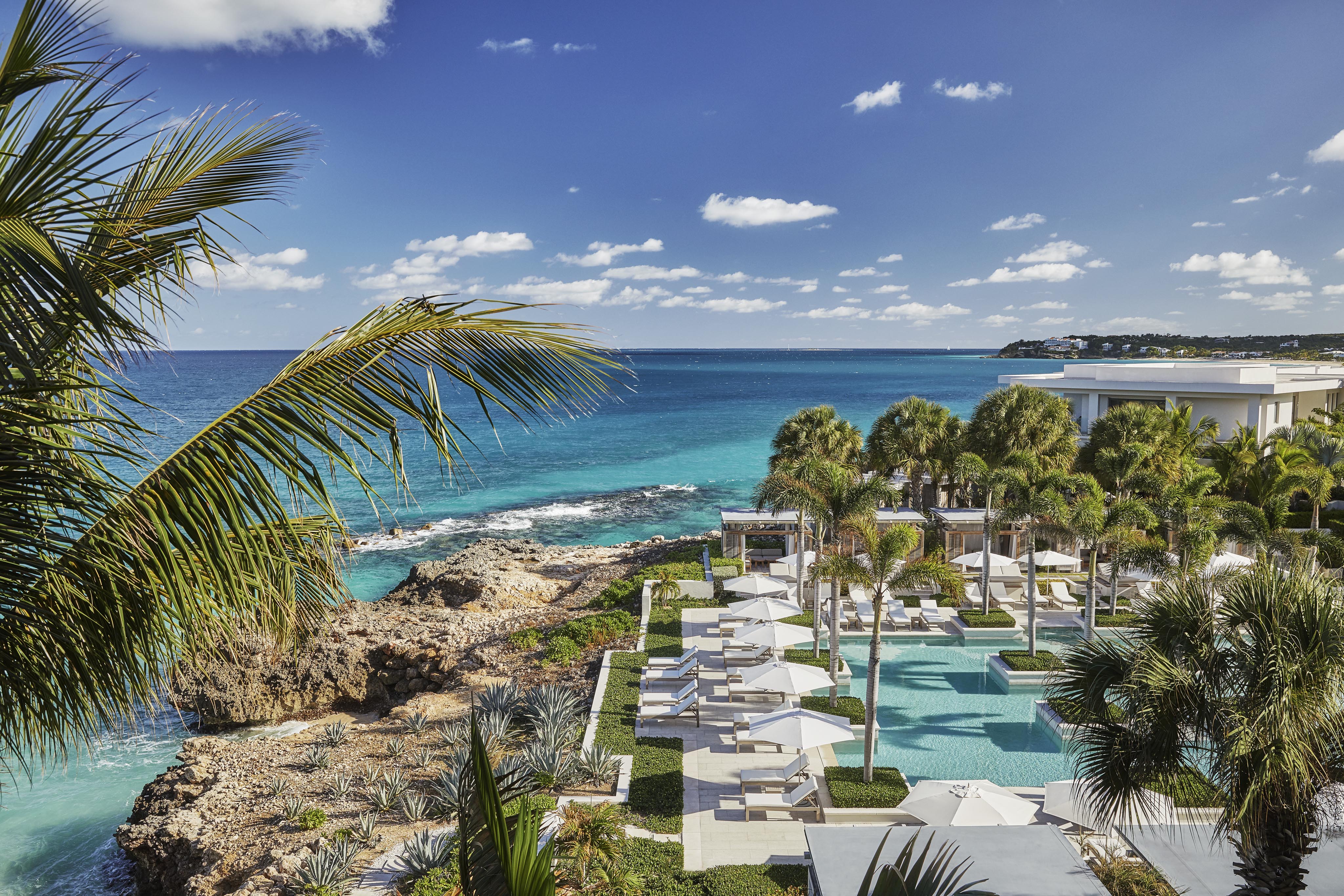Anguilla Four Seasons resort pool area with lounge chairs and umbrellas overlooks turquoise ocean water under a clear blue sky.