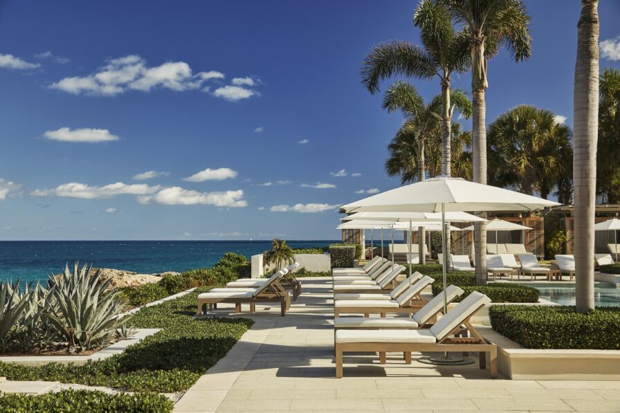 Poolside lounge chairs and Four Seasons Anguilla resort umbrellas overlooking a clear blue ocean. Palm trees and greenery surround the area under a bright, partly cloudy sky.