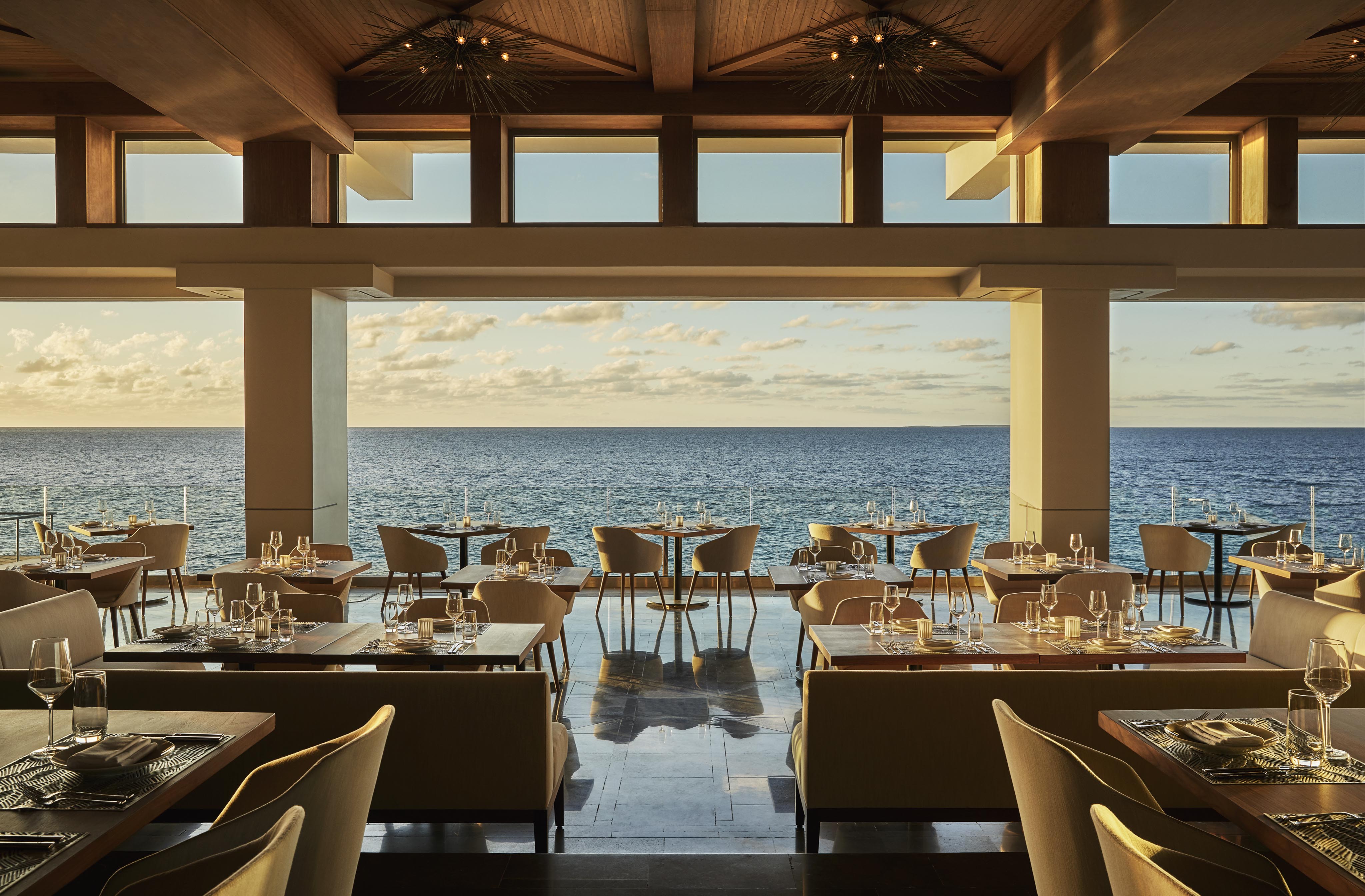 Four Seasons Anguilla resort interior with modern furniture and elegant table settings, featuring large windows offering a panoramic ocean view under a partly cloudy sky.