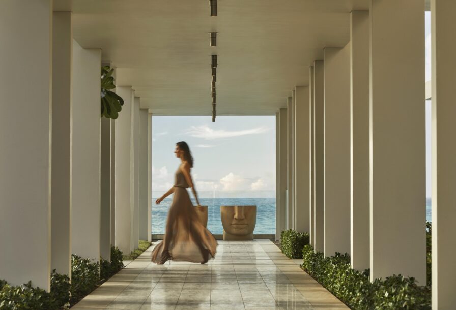 A person in a flowing dress walks down a corridor with white pillars, leading to a large, face-like sculpture. Ocean view is visible in the background.